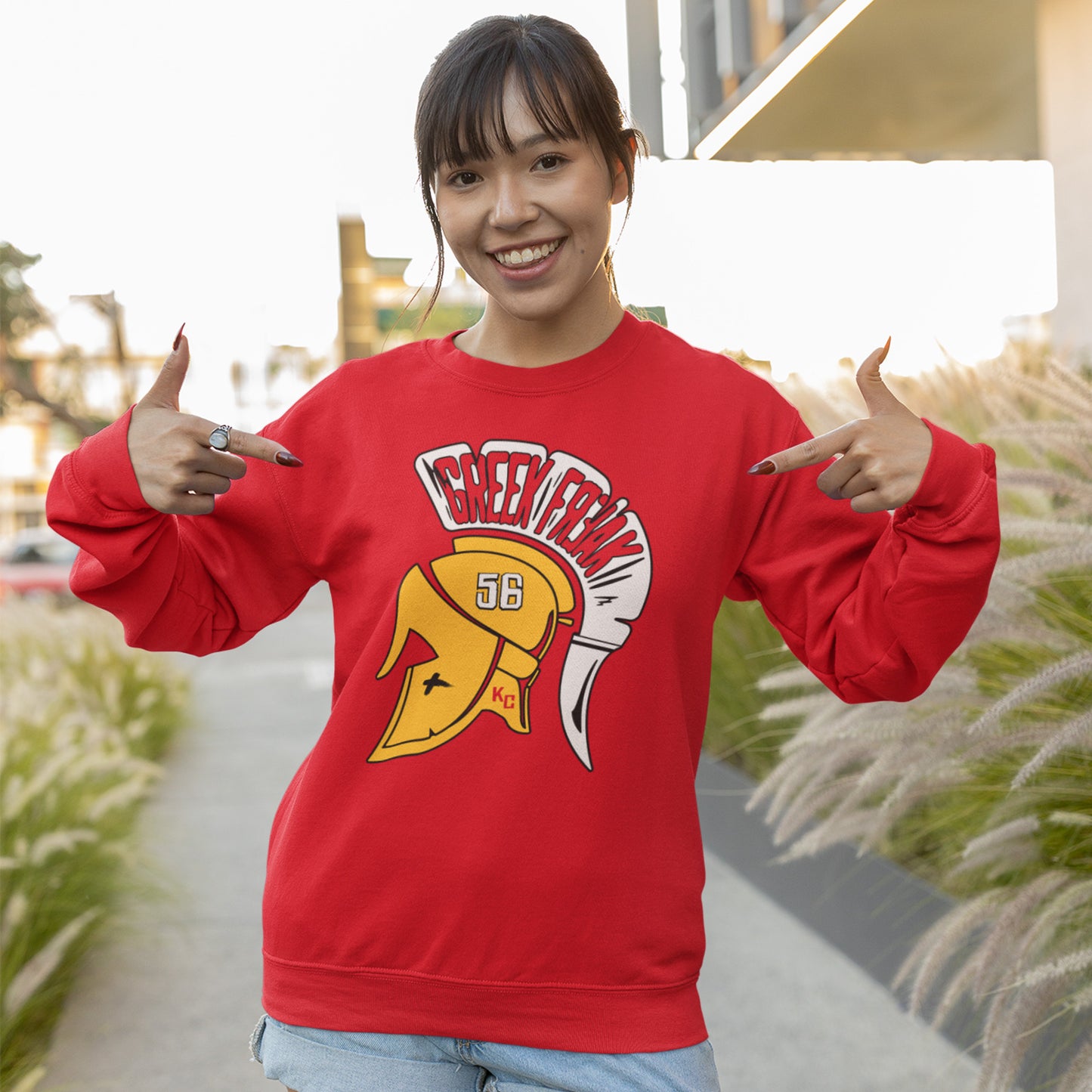 KC Swag Kansas City Chiefs White & Gold Greek Freak (with Trojan Helmet) on a Red Crewneck Sweatshirt worn by a smiling female model pointing at her chest while standing on an apartment building sidewalk