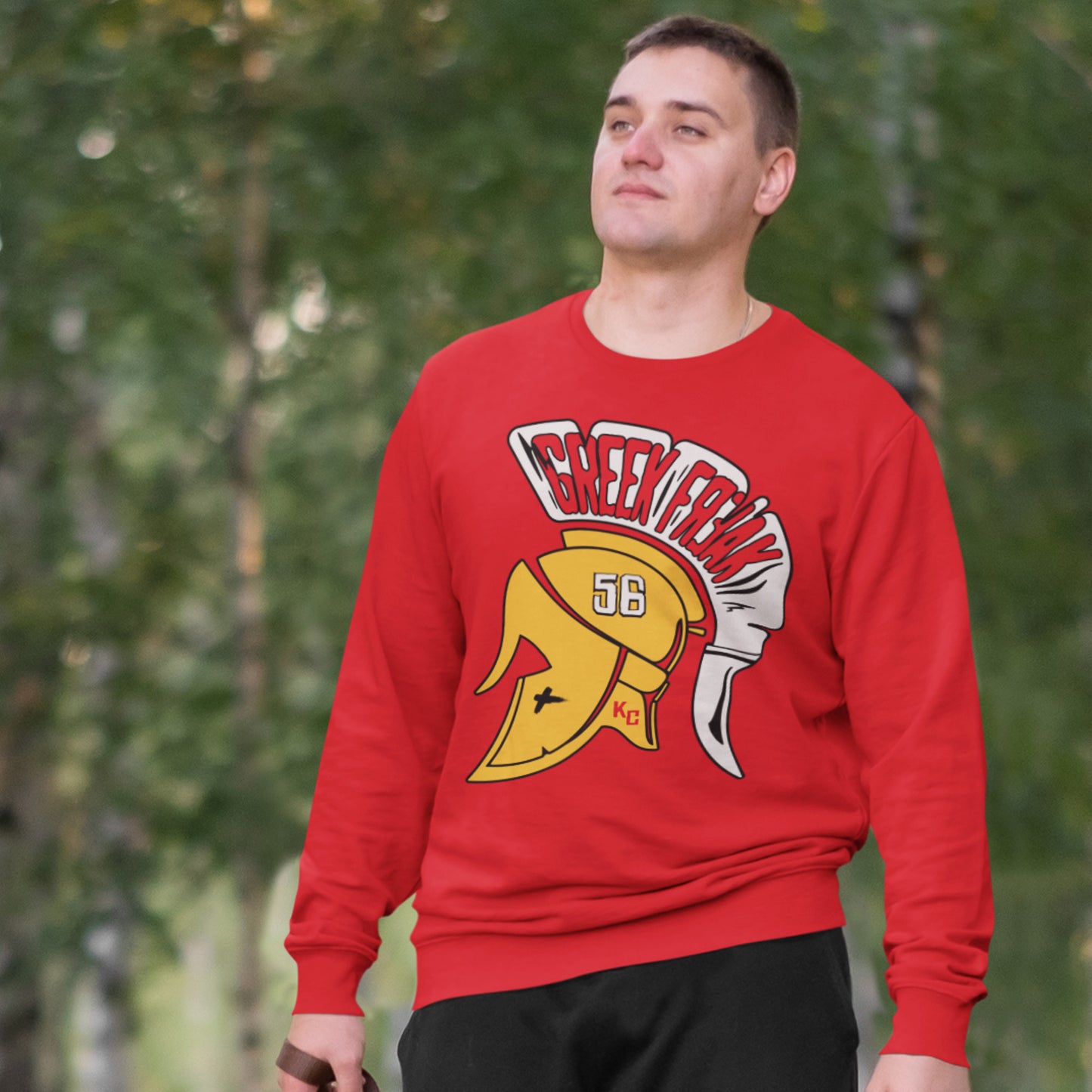 KC Swag Kansas City Chiefs White & Gold Greek Freak (with Trojan Helmet) on a Red Crewneck Sweatshirt worn by male model standing on tree-lined sidewalk