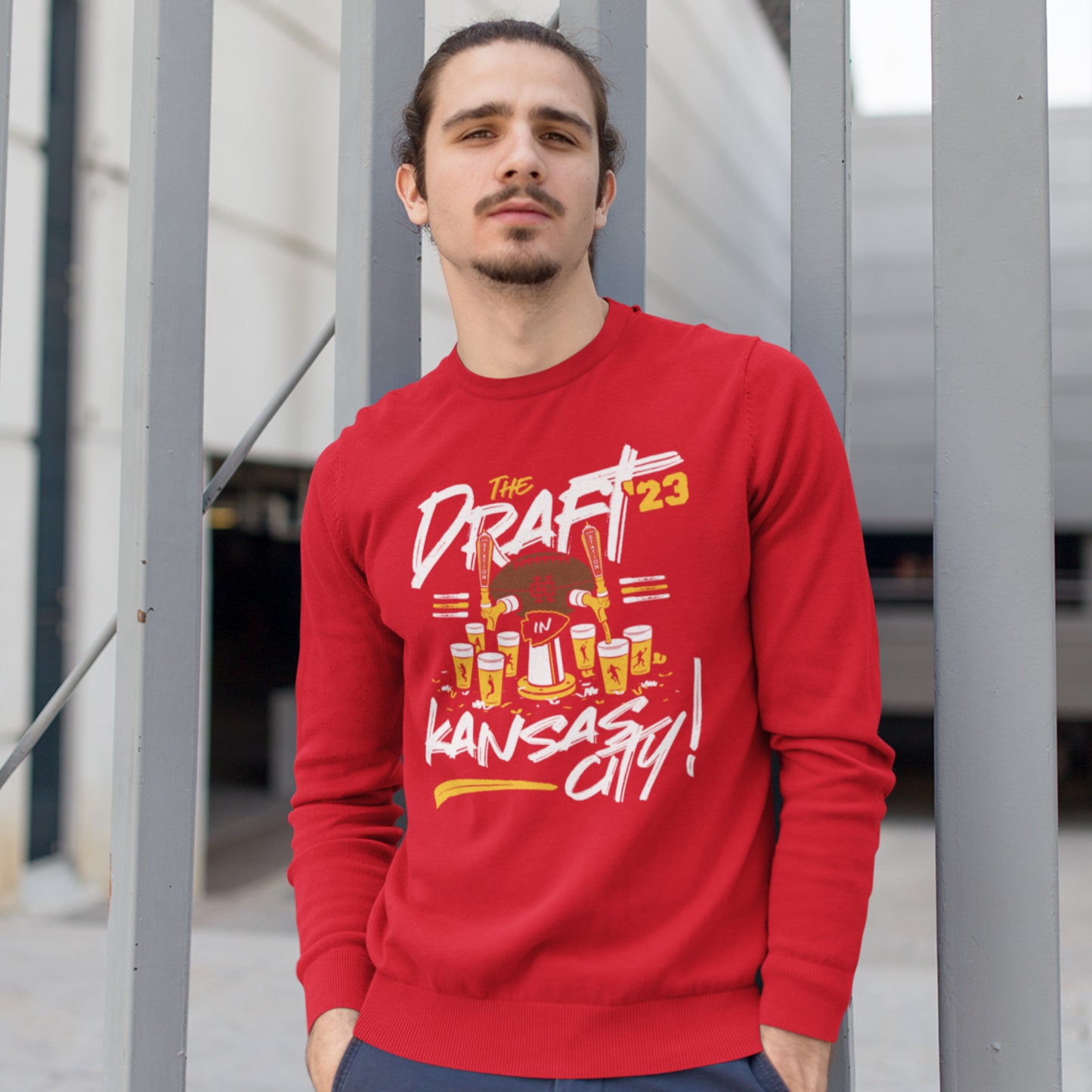 KC Swag Kansas City Chiefs Distressed White & Gold Draftin' IN KC 2023 on a Red Crewneck Sweatshirt worn by male model with a goatee leaning against a large metal gate in an urban setting