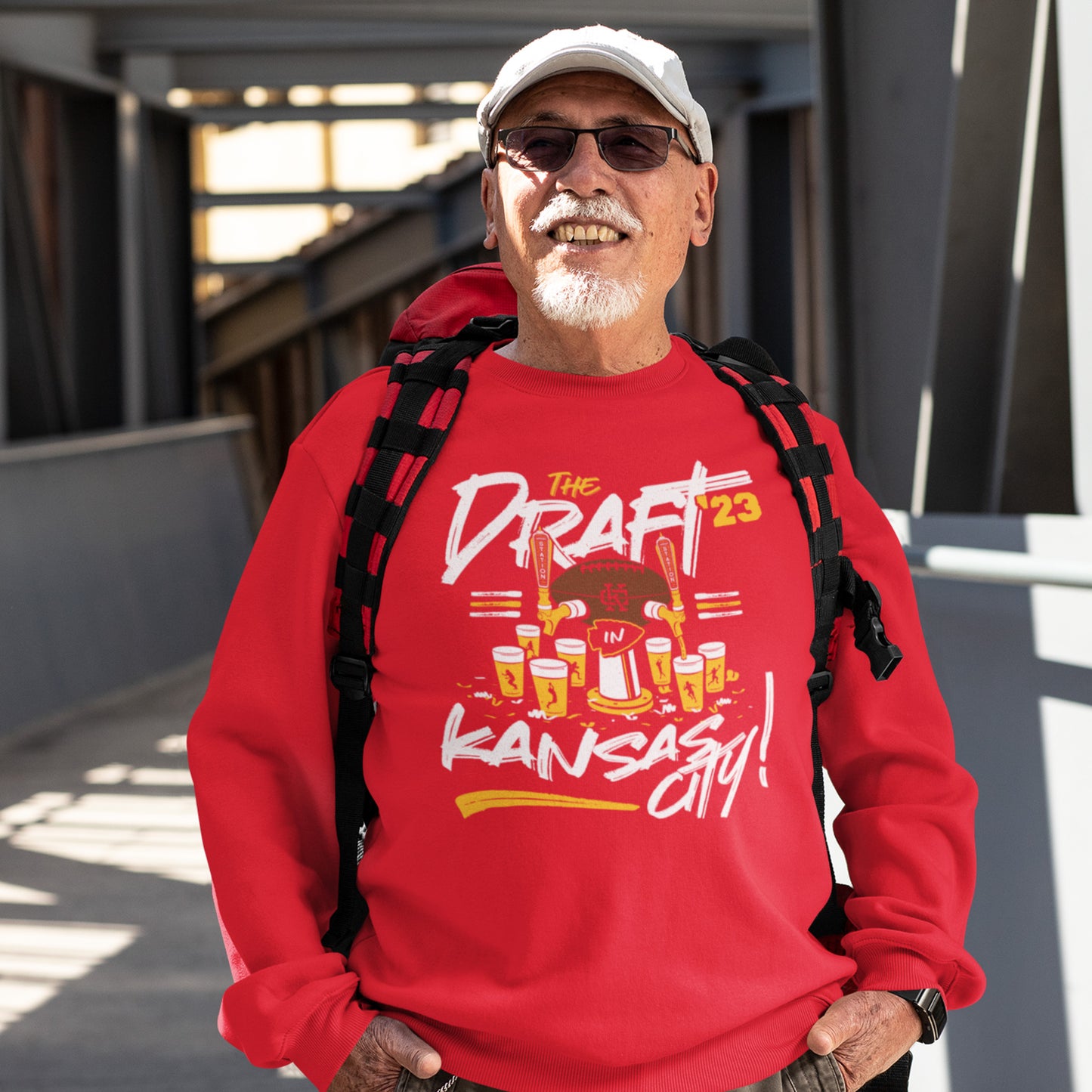 KC Swag Kansas City Chiefs Distressed White & Gold Draftin' IN KC 2023 on a Red Crewneck Sweatshirt worn by an older male model wearing a hat and backpack in an urban setting