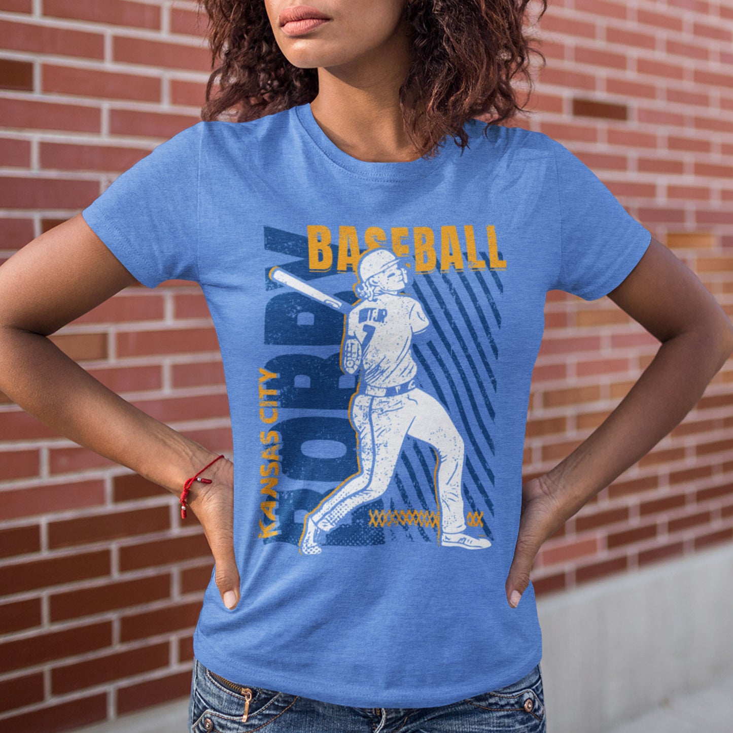 kc swag, kansas city royals, heather columbia blue unisex t-shirt featuring a white image of a baseball player, Bobby Witt Jr., along with gold and royal blue graphics saying, "Bobby Baseball" and "Kansas City" in the style of a distressed retro design, worn by a curly-hair female model standing in front of a brick wall