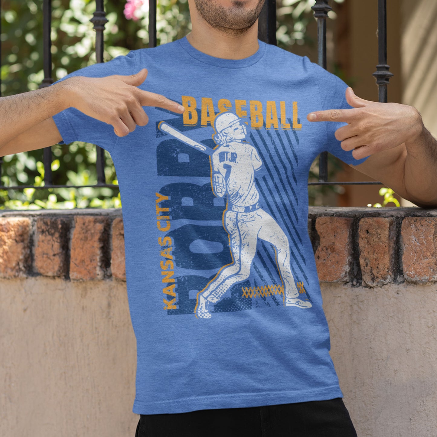 kc swag, kansas city royals, heather columbia blue unisex t-shirt featuring a white image of a baseball player, Bobby Witt Jr., along with gold and royal blue graphics saying, "Bobby Baseball" and "Kansas City" in the style of a distressed retro design, worn by a male model showing off his shirt by pointing at his chest
