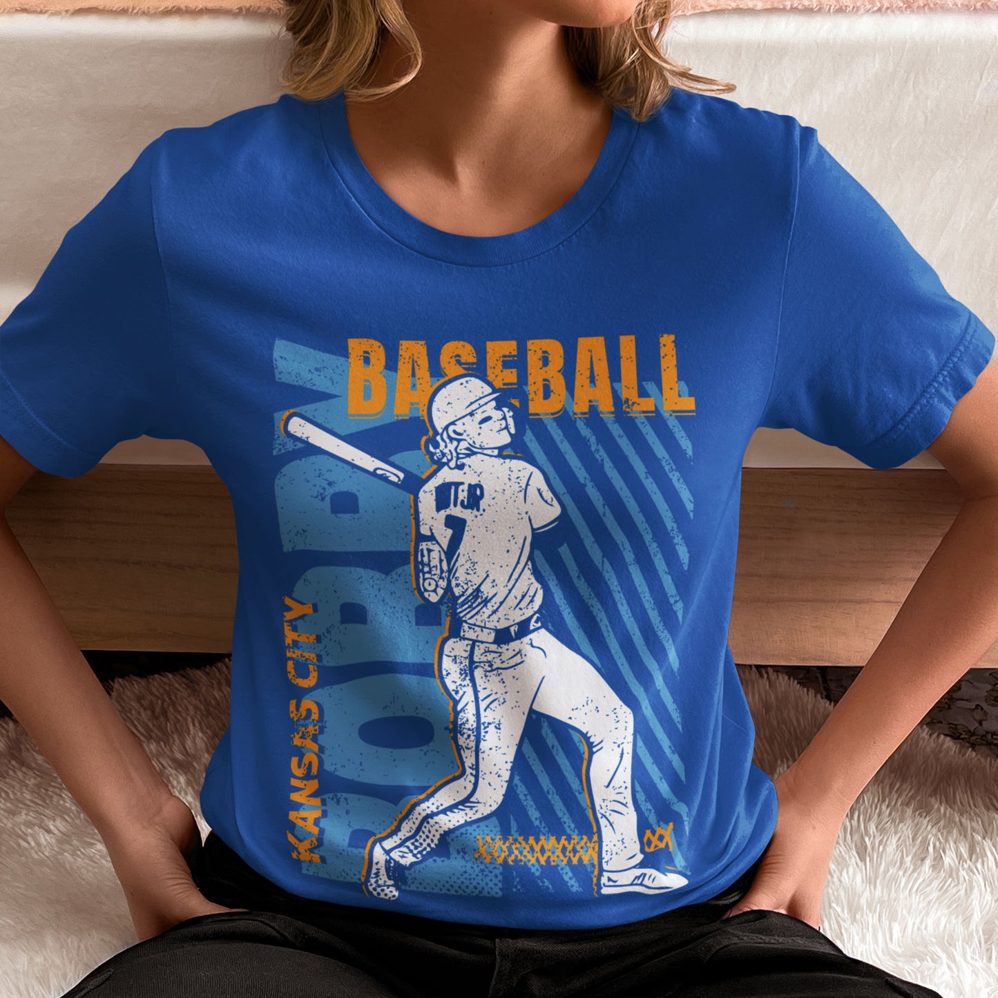 kc swag, kansas city royals, royal blue unisex t-shirt featuring a white image of a baseball player, Bobby Witt Jr., along with gold and light blue graphics saying, "Bobby Baseball" and "Kansas City" in the style of a distressed retro design, worn by female model sitting on the floor of a bedroom with her hands on her hips