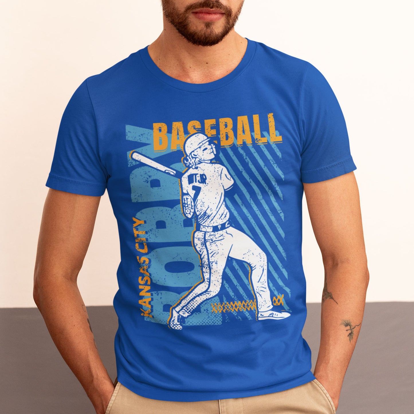 kc swag, kansas city royals, royal blue unisex t-shirt featuring a white image of a baseball player, Bobby Witt Jr., along with gold and light blue graphics saying, "Bobby Baseball" and "Kansas City" in the style of a distressed retro design, worn by a bearded male model standing in front of a two-tone grey studio wall