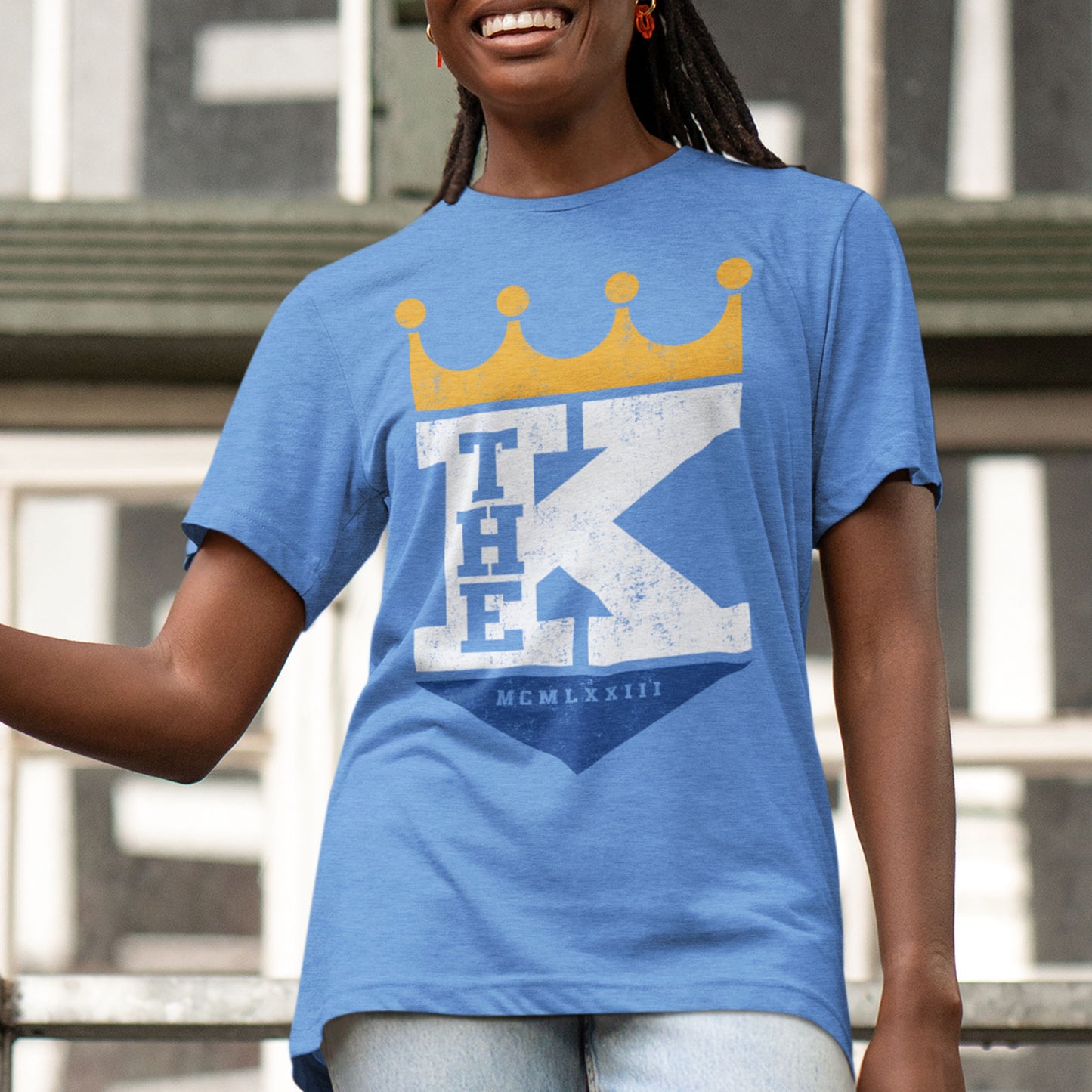 kc swag, kansas city royals, heather columbia blue unisex t-shirt featuring a bold white text that says, "The K" sandwiched with a distressed, worn-out look, between a gold crown on top and a royal blue triangle underneath forming a silhouette resembling the team logo, worn by a female model with raids standing on an outdoor metal staircase