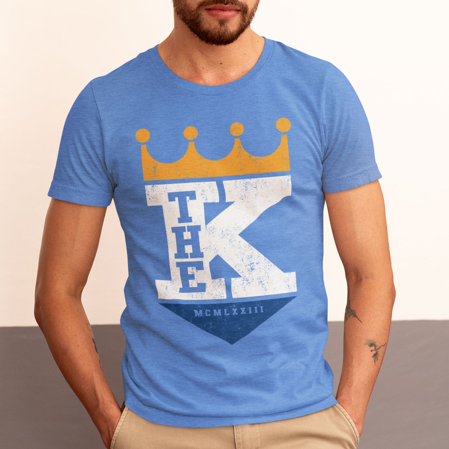 kc swag, kansas city royals, heather columbia blue unisex t-shirt featuring a bold white text that says, "The K" sandwiched with a distressed, worn-out look, between a gold crown on top and a royal blue triangle underneath forming a silhouette resembling the team logo, worn by a bearded man standing in front of a two-tone grey studio wall