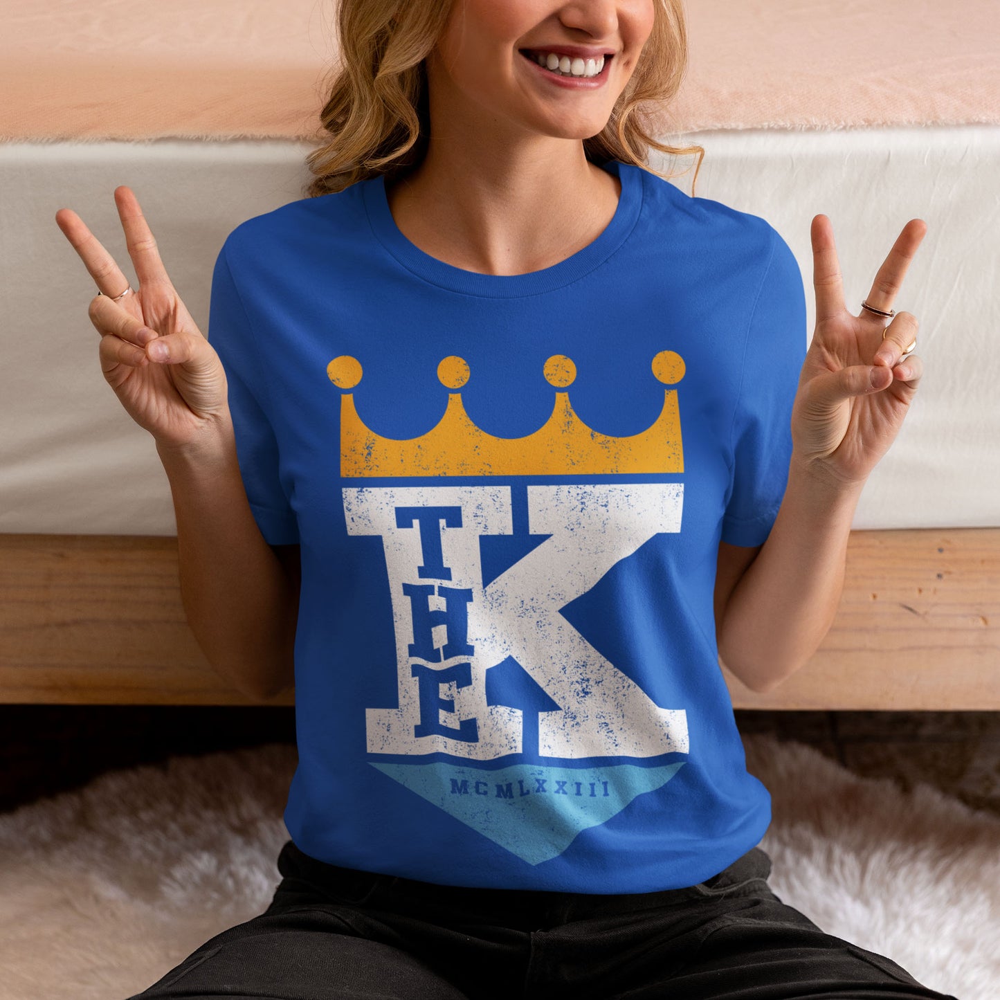kc swag, kansas city royals, royal blue unisex t-shirt featuring a bold white text that says, "The K" sandwiched with a distressed, worn-out look, between a gold crown on top and a light blue triangle underneath forming a silhouette resembling the team logo, worn by a female model sitting on the floor of a bedroom flashing peace signs