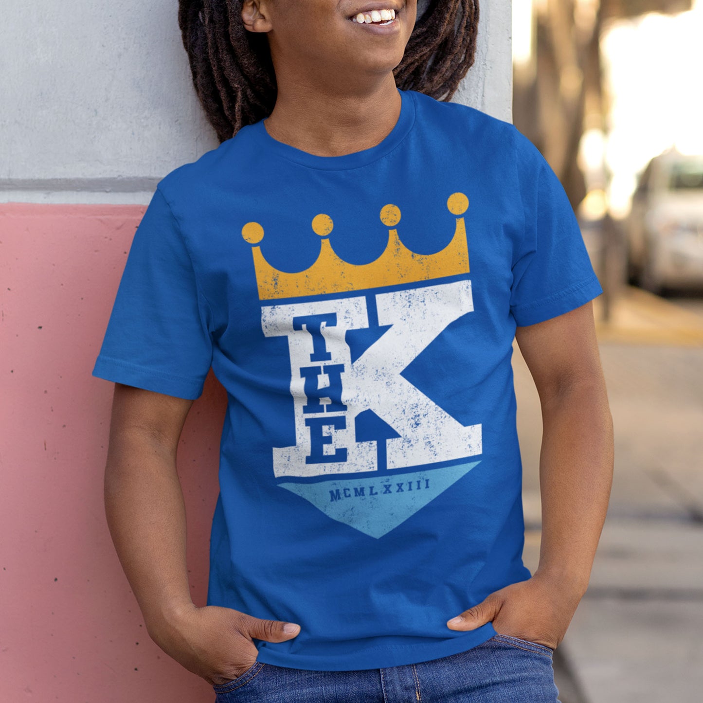 kc swag, kansas city royals, royal blue unisex t-shirt featuring a bold white text that says, "The K" sandwiched with a distressed, worn-out look, between a gold crown on top and a light blue triangle underneath forming a silhouette resembling the team logo, worn by a laughing male model with dreadlocks leaning on a two-tone concrete wall