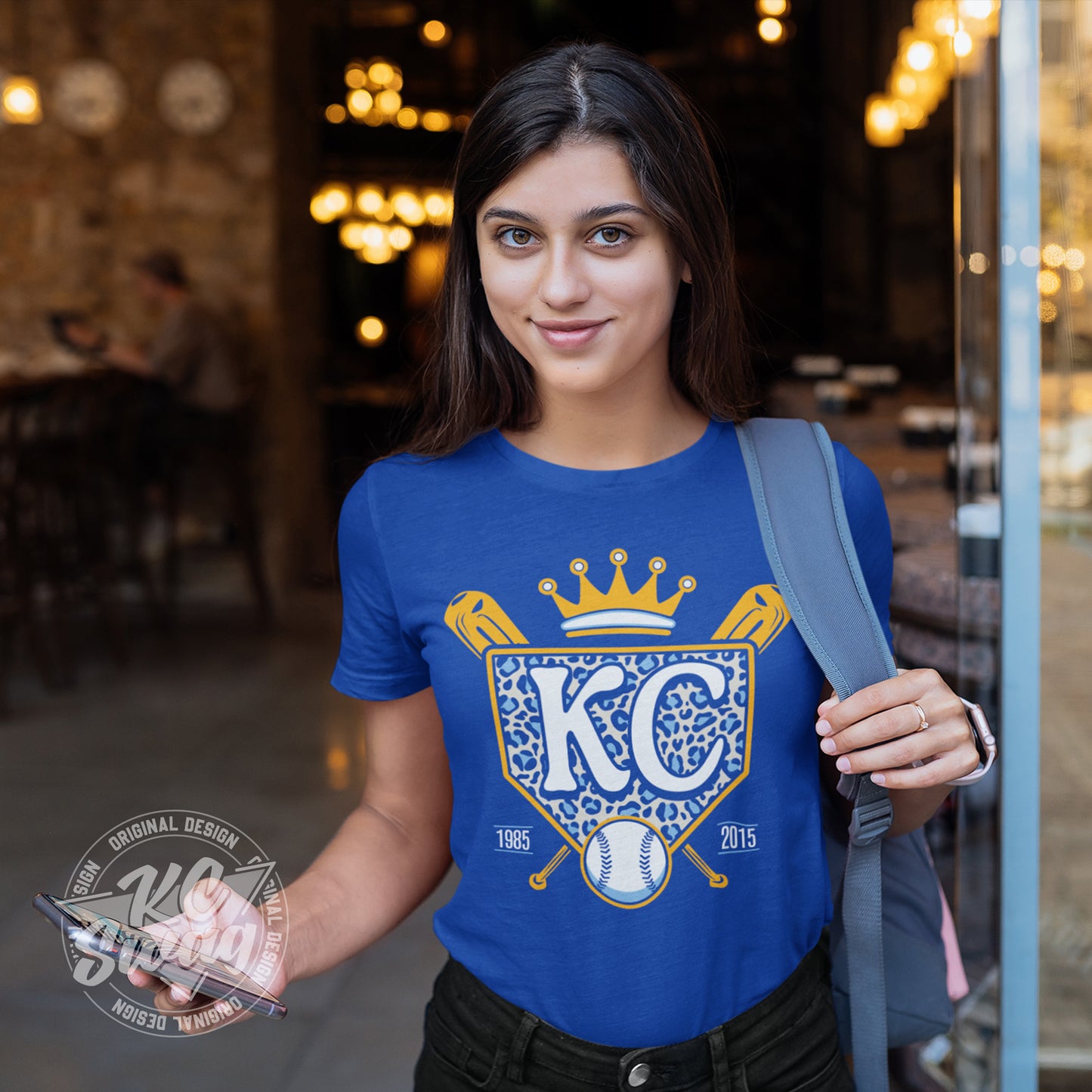 KC Swag - Kansas City Royals, Cheetah Home Plate design on royal blue unisex t-shirt worn by female model standing in cafe doorway holding her smart phone and backpack