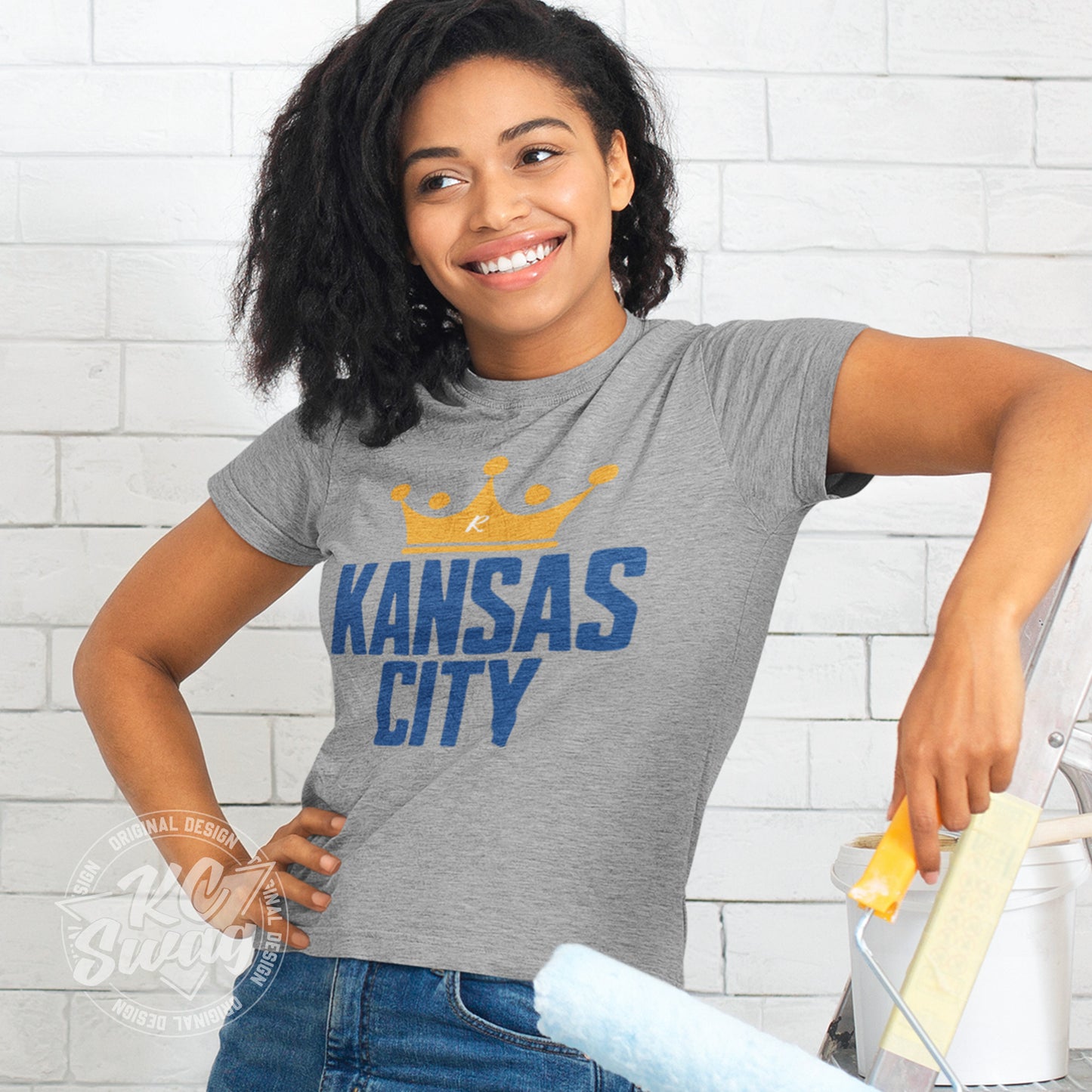 KC Swag - Kansas City Royals, Crowned KC design on athletic heather grey unisex t-shirt worn by female model leaning on a ladder and holding a paint roller in front of a white brick wall