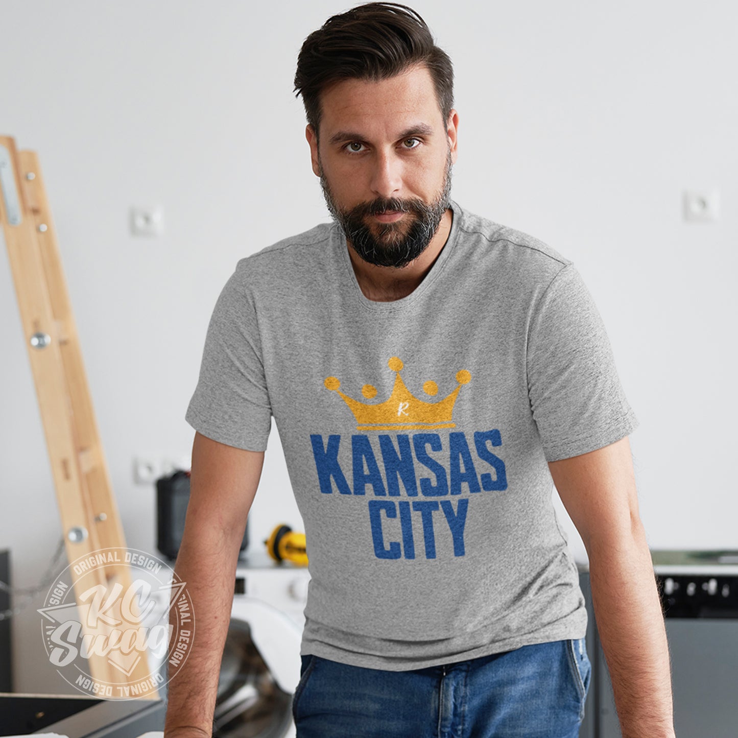 KC Swag - Kansas City Royals, Crowned KC design on athletic heather grey unisex t-shirt worn by male model leaning on table in a home construction zone