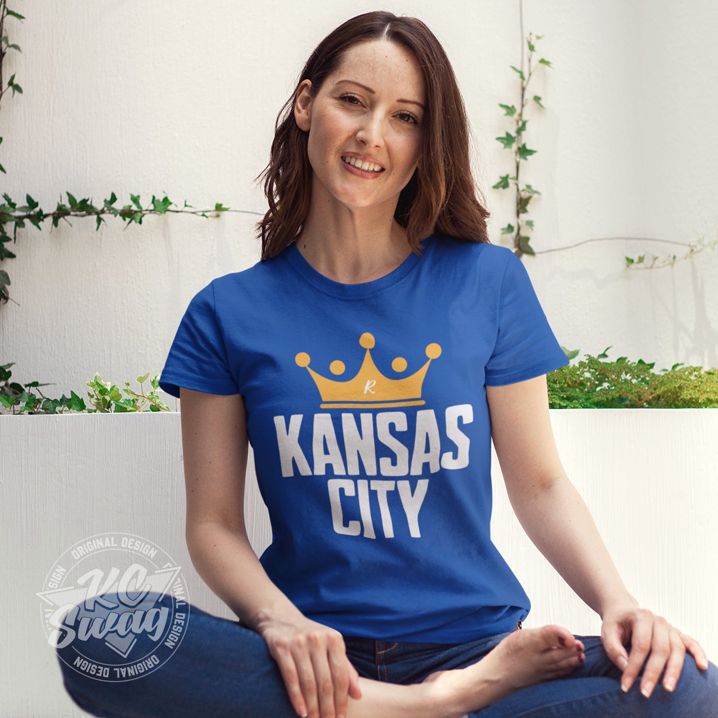 KC Swag - Kansas City Royals, Crowned KC design on royal blue unisex t-shirt worn by female model sitting on the floor in front of a concrete wall
