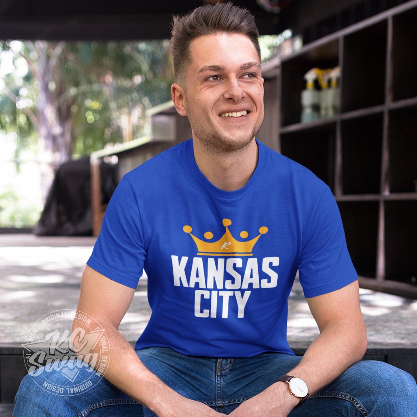 KC Swag - Kansas City Royals, Crowned KC design on royal blue unisex t-shirt worn by male model sitting on steps in front of a rustic barn setting