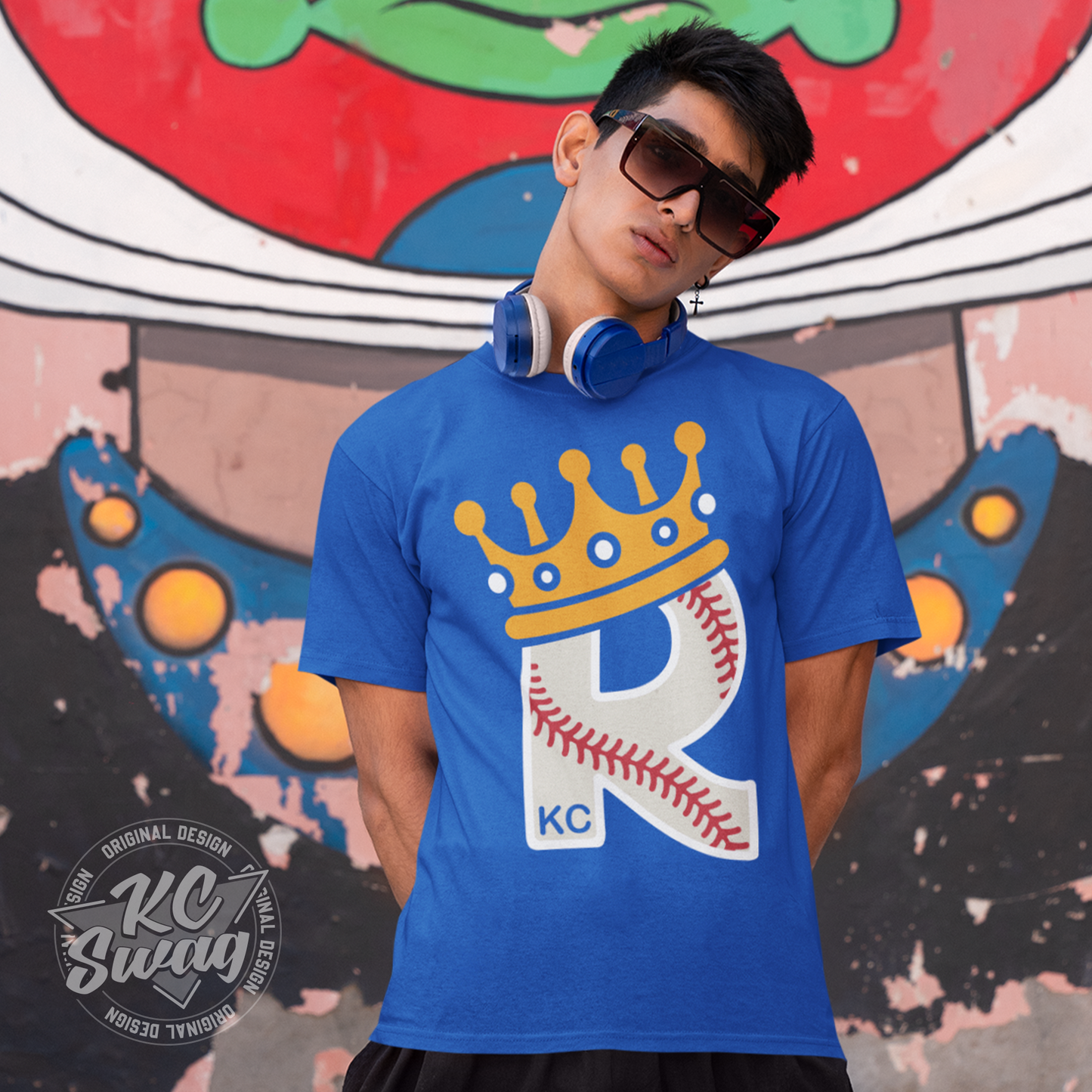 KC Swag - Kansas City Royals CROWN R-BALL KC on true royal blue unisex t-shirt worn by male model standing in front of a brightly colored graffiti wall with headphones around his neck