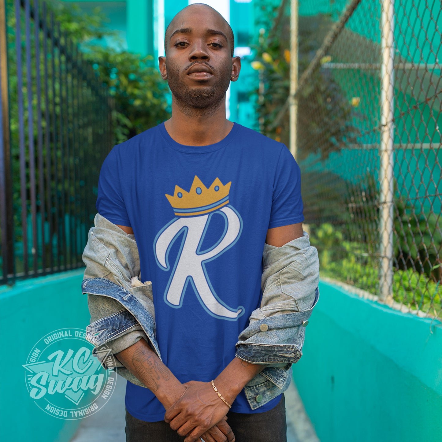 KC Swag - Kansas City Royals, Crown R design on royal blue unisex t-shirt worn by male model standing on a cement walkway in front of a bright teal building