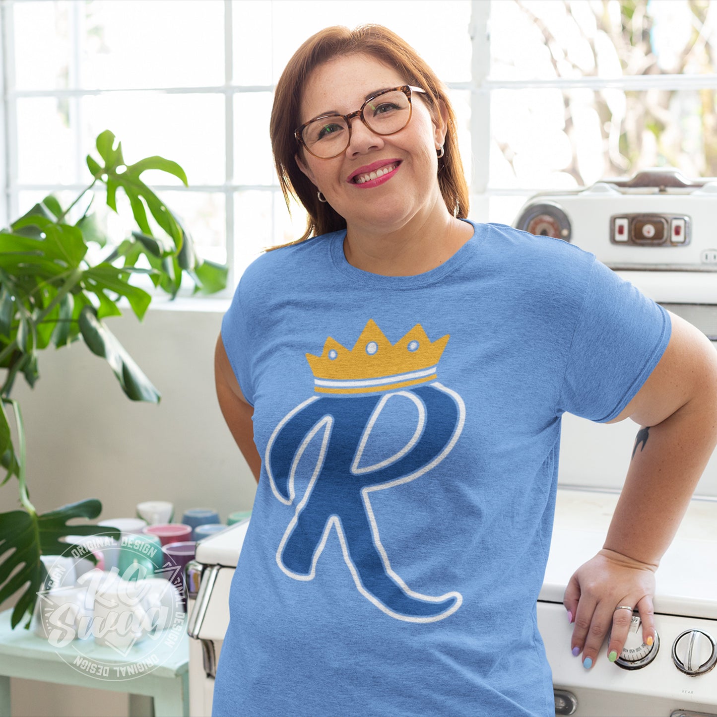 KC Swag - Kansas City Royals, Crown R design on heather columbia blue unisex t-shirt worn by female model leaning on vintage store in bright kitchen
