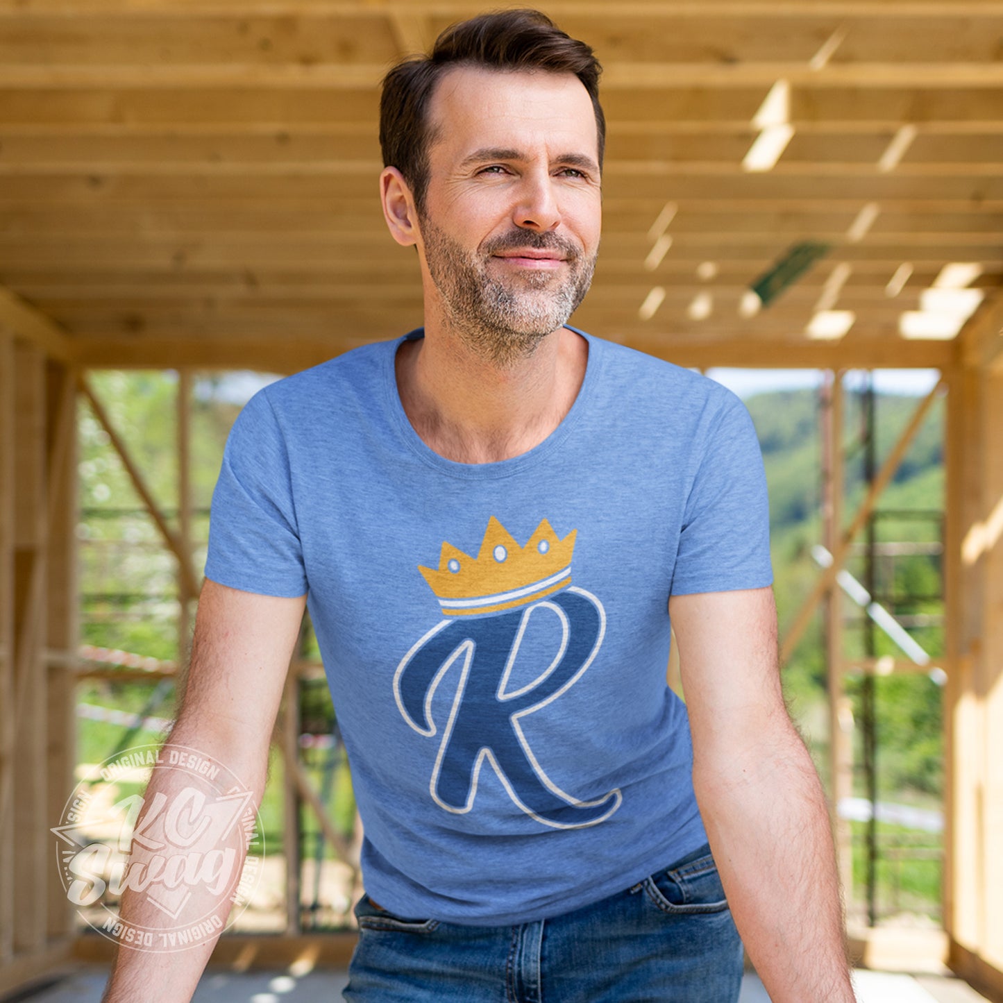 KC Swag - Kansas City Royals, Crown R design on heather columbia blue unisex t-shirt worn by male model leaning on open window frame in an unfinished home construction zone