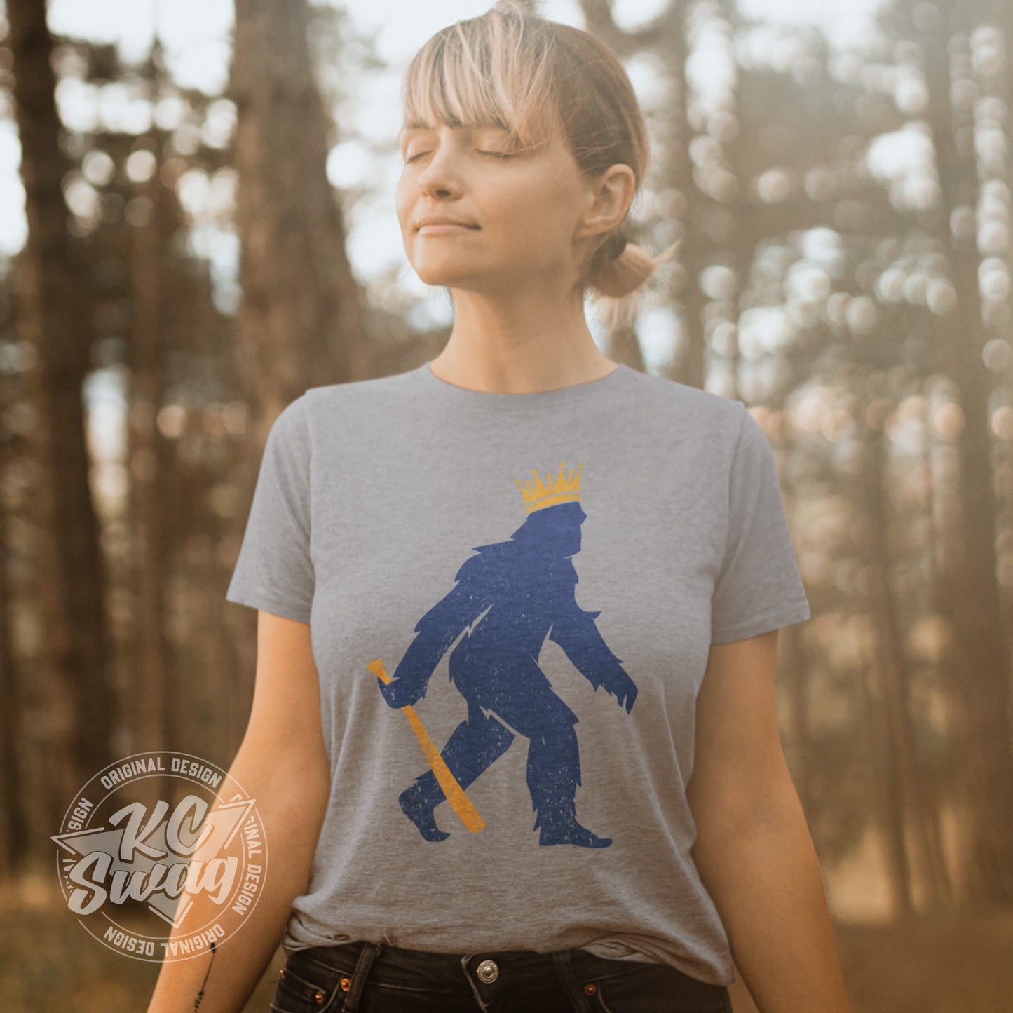 KC Swag - Kansas City Royals, Crowned Pasquatch design on athletic heather grey unisex t-shirt worn by female model basking in the sunlight in a forest setting