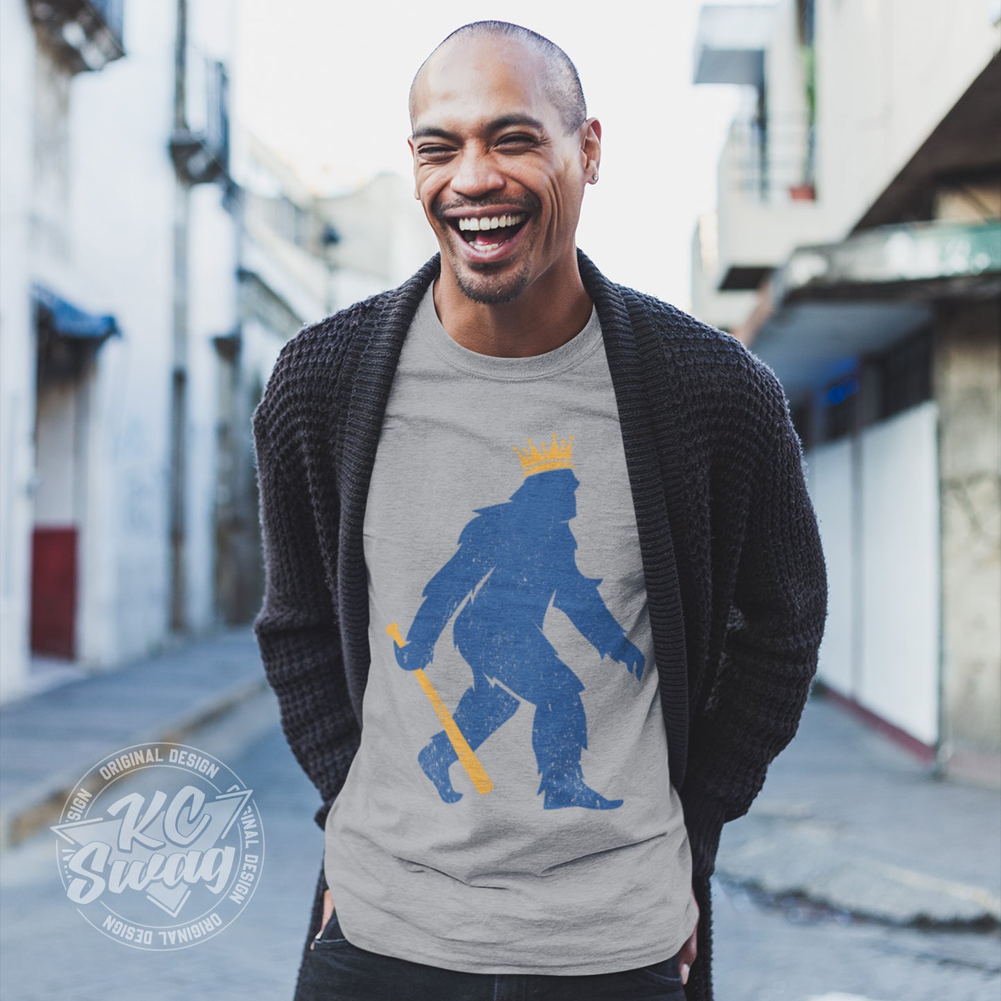 KC Swag - Kansas City Royals, Crowned Pasquatch design on athletic heather grey unisex t-shirt worn by laughing male model under open sweater in an urban alley