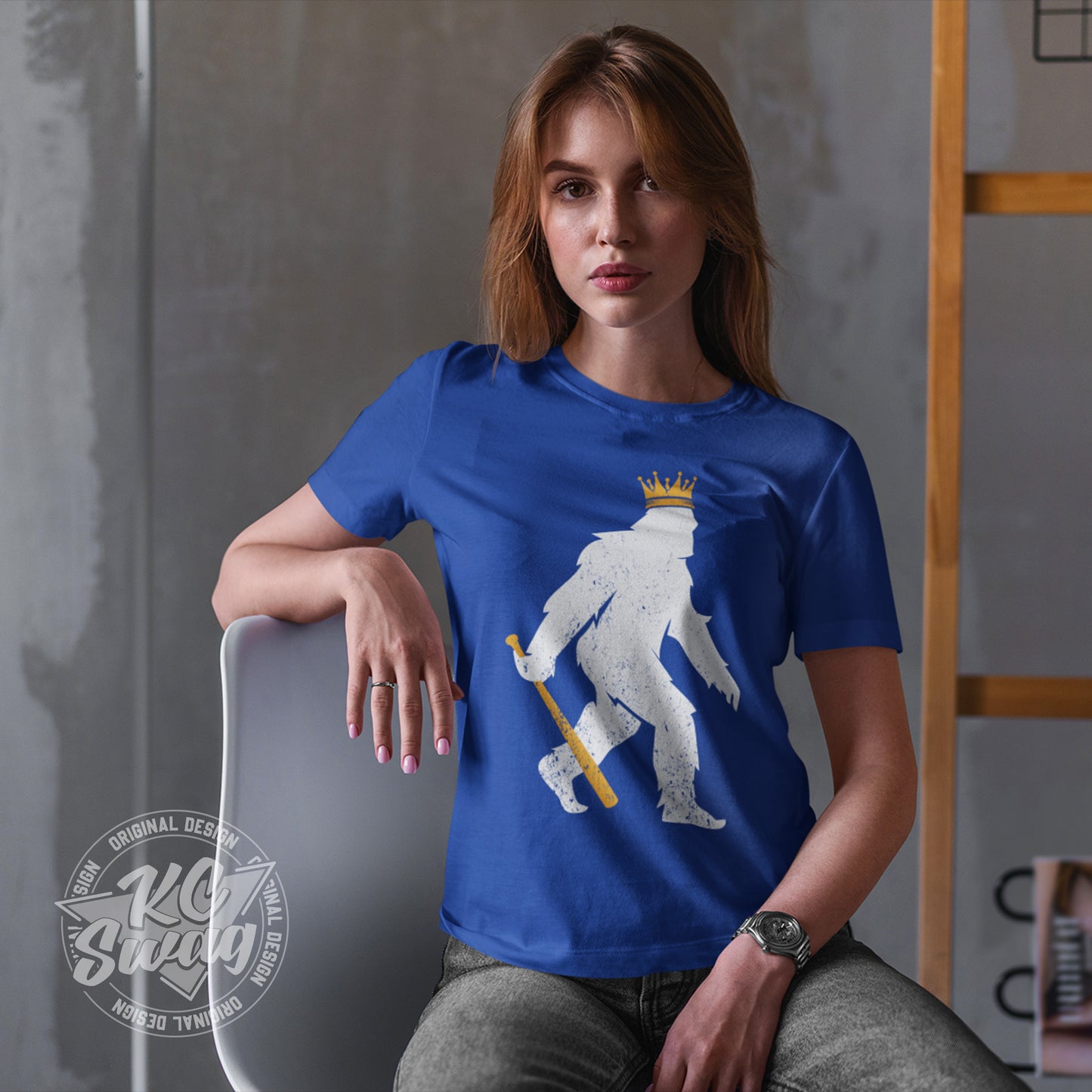 KC Swag - Kansas City Royals, Crowned Pasquatch design on royal blue unisex t-shirt worn by female model stting in a chair next to a clothing rack in a studio