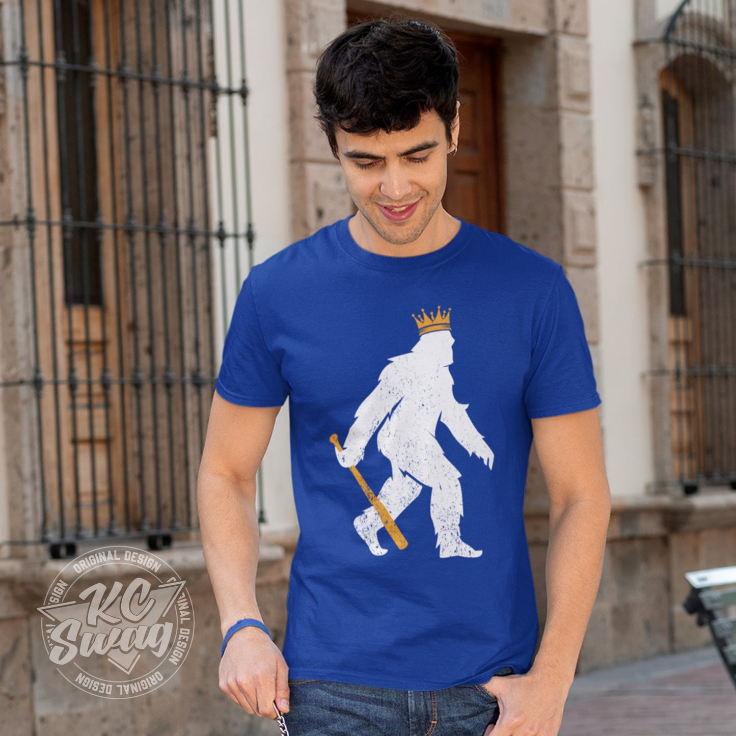 KC Swag - Kansas City Royals, Crowned Pasquatch design on royal blue unisex t-shirt worn by male model walking in an open-air plaza