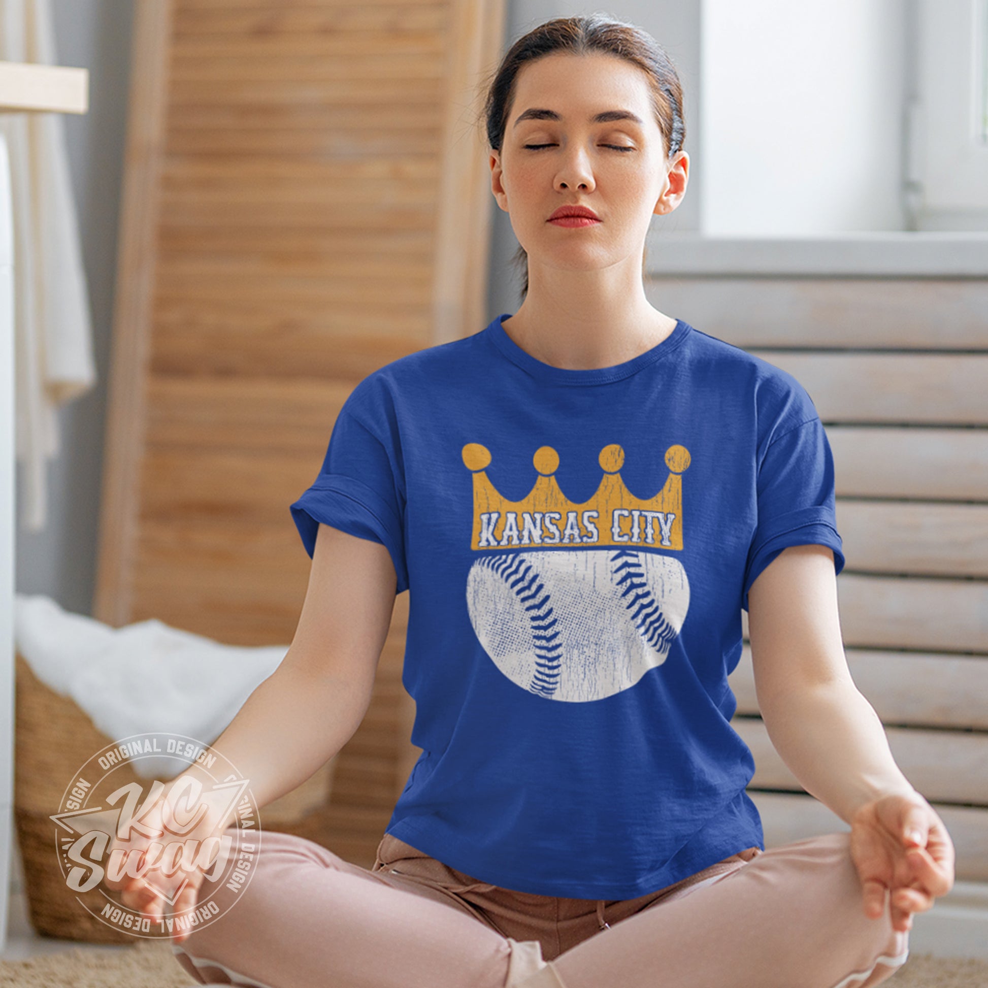 KC Swag - Kansas City Royals, KC Baseball Crown design on royal blue unisex t-shirt worn by female model sitting cross-legged on the floor meditating