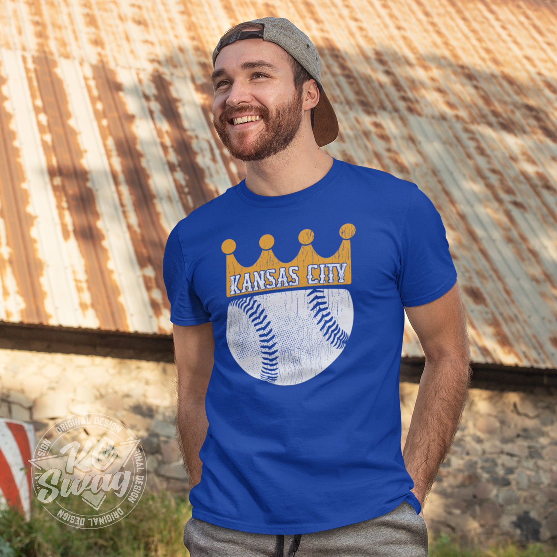 KC Swag - Kansas City Royals, KC Baseball Crown design on royal blue unisex t-shirt worn by male model standing in front of a rustic old barn on a farm