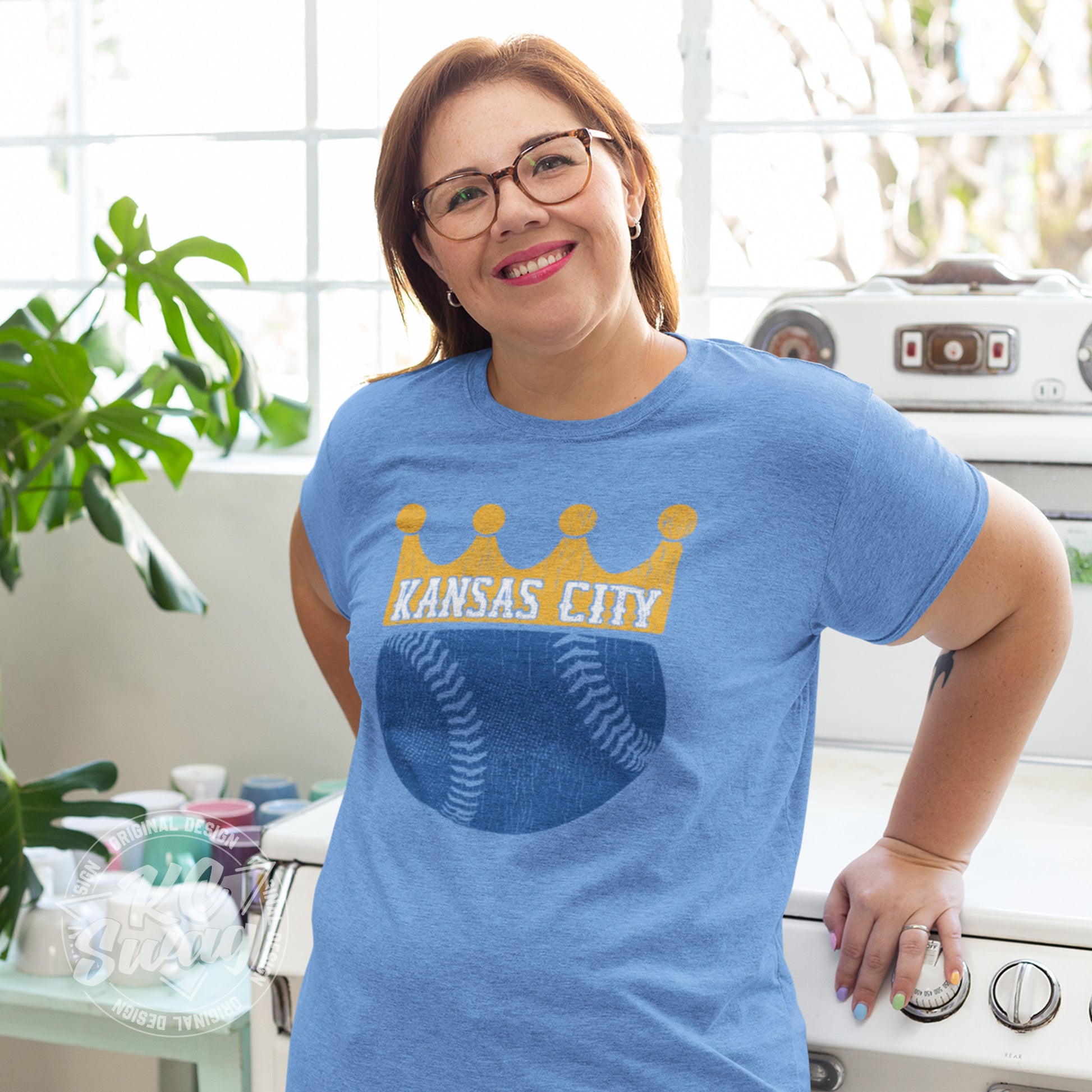 KC Swag - Kansas City Royals, KC Baseball Crown design on heather columbia blue unisex t-shirt worn by female model leaning on vintage stove in a bright kitchen