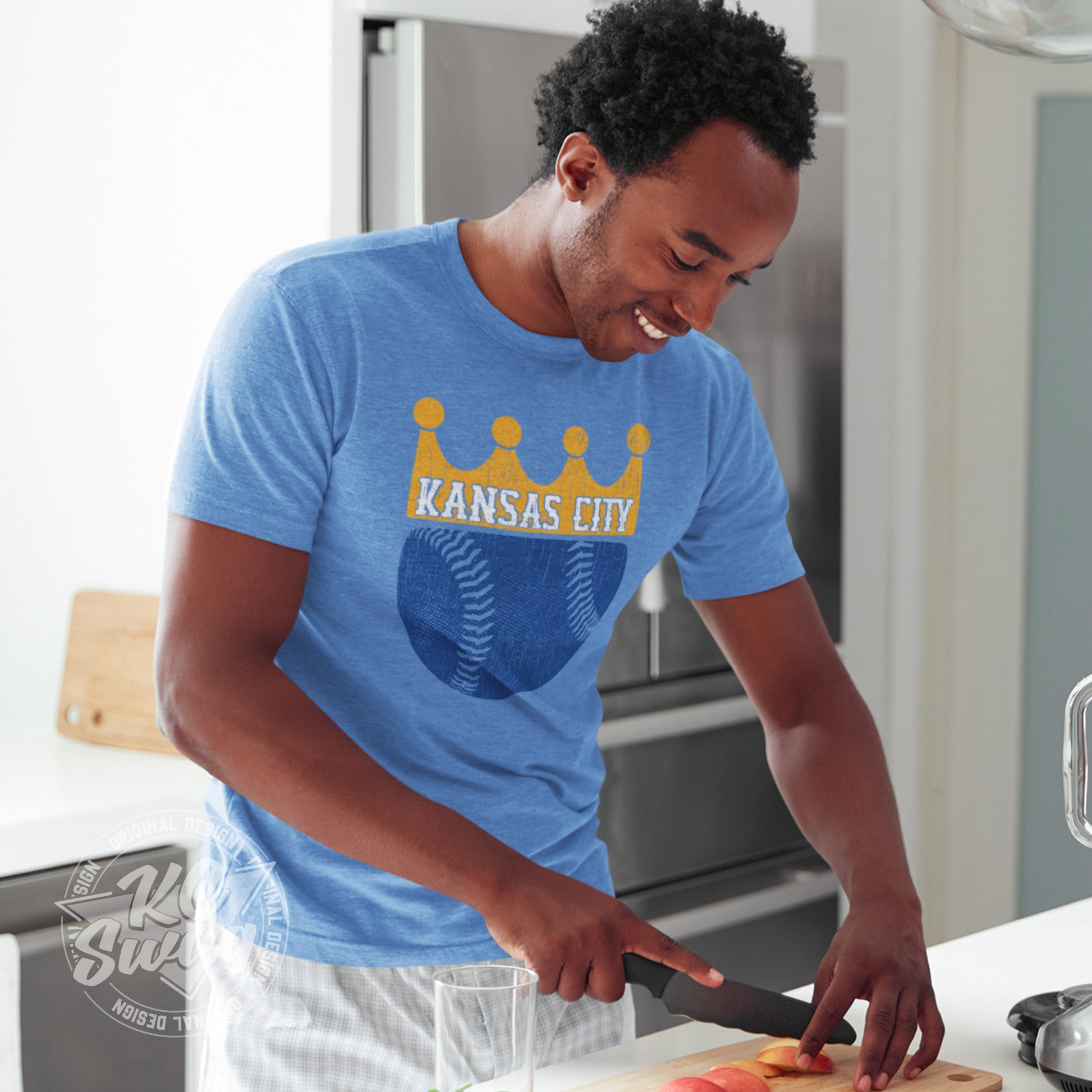 KC Swag - Kansas City Royals, KC Baseball Crown design on heather columbia blue unisex t-shirt worn by male model slicing fruit for a smoothie in his kitchen