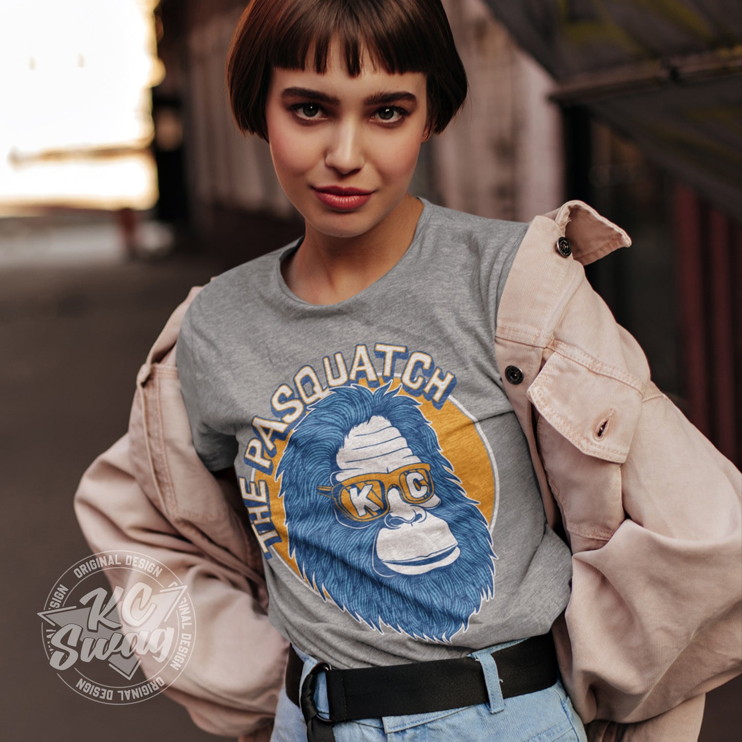 KC Swag - Kansas City Royals, Shaded KC Pasquatch on athletic heather grey unisex t-shirt worn by female model under open jack standing in front of a rustic barn setting