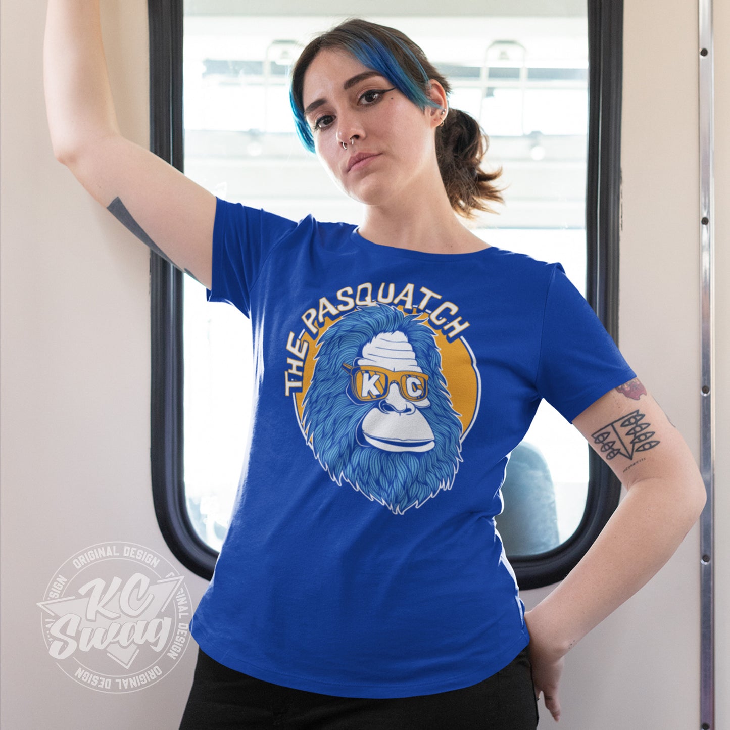 KC Swag - Kansas City Royals, Shaded KC Pasquatch on royal blue unisex t-shirt worn by female model standing in front of window on a city streetcar