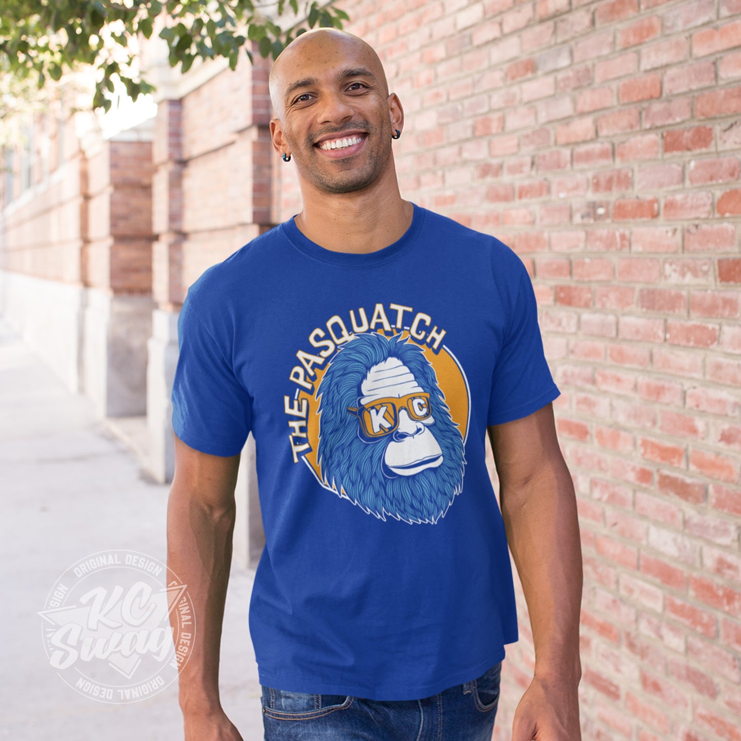 KC Swag - Kansas City Royals, Shaded KC Pasquatch on royal blue unisex t-shirt worn by male model walking on sidewalk next to brick wall