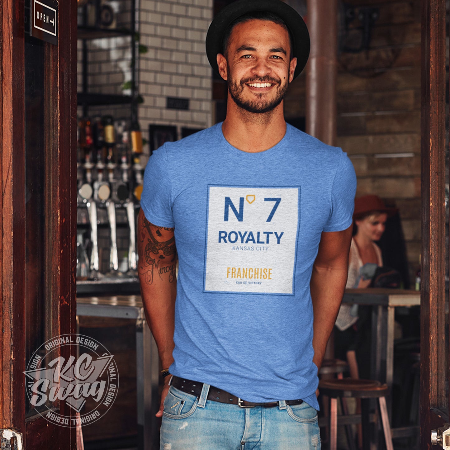 KC Swag - Kansas City Royals, Royalty Number 7 Franchise design on heather columbia blue unisex t-shirt worn by male model wearing a brimed hat standing in the doorway to a retro cafe