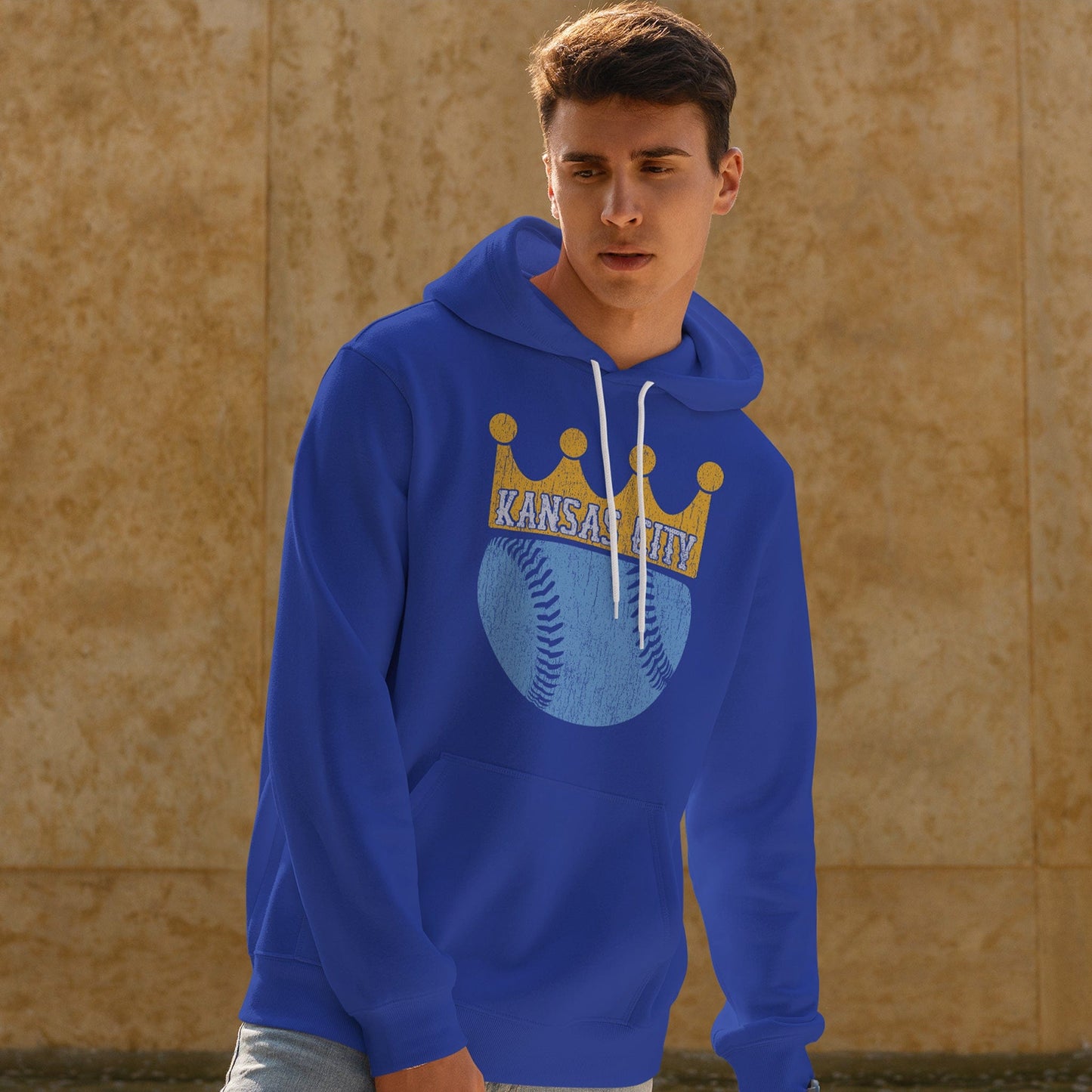 KC Swag Kansas City Royals powder blue baseball wearing a gold crown with KANSAS CITY text on royal blue pull-over hoodie worn by male model in front of stone wall