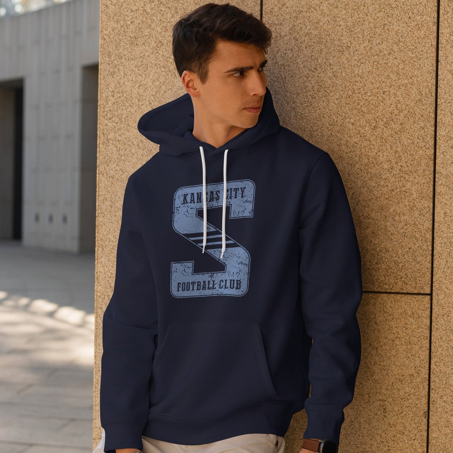 KC Swag Sporting Kansas City powder blue KANSAS CITY FOOTBALL CLUB in STRIPED S on navy fleece pull-over hoodie worn by male model leaning against stone wall in outdoor plaza