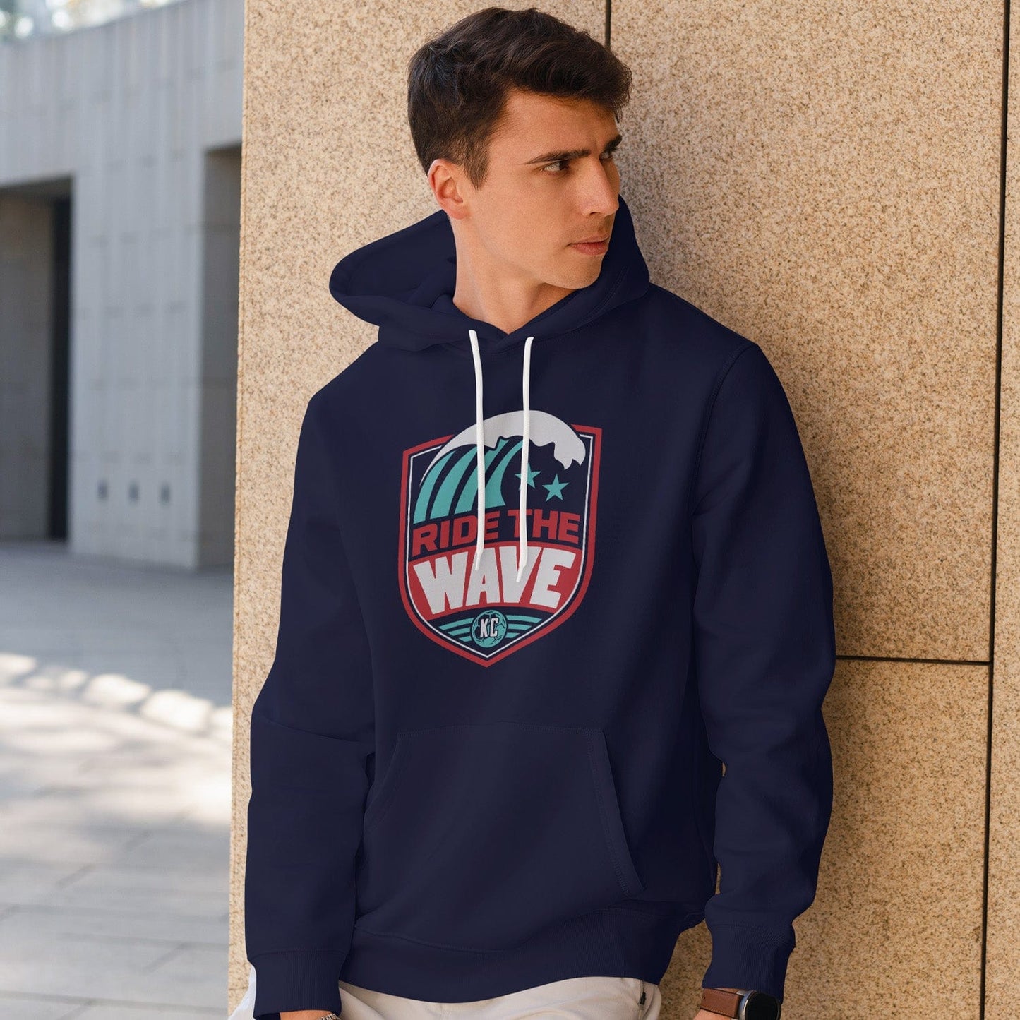 KC Swag Kansas City Current RIDE THE WAVE on navy fleece pullover hoodie worn by male model standing near stone wall in outdoor plaza