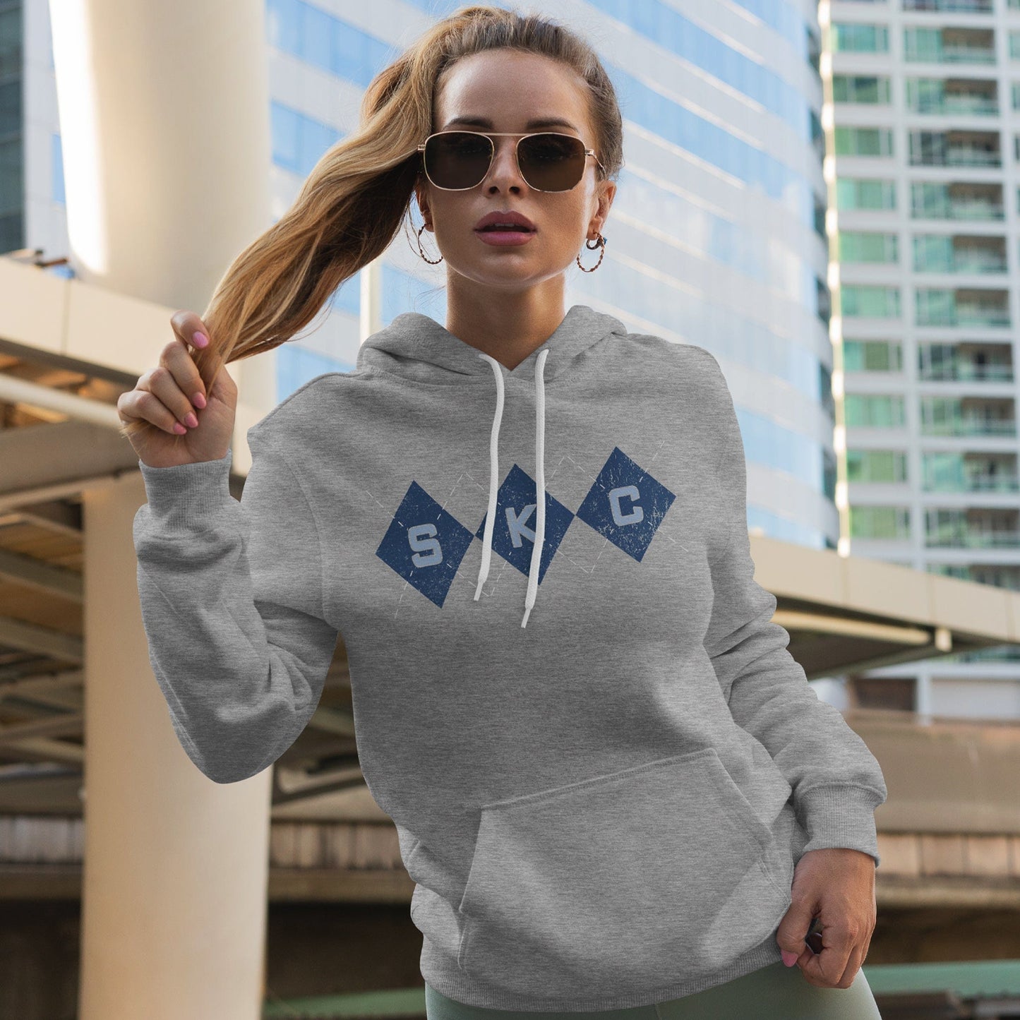 KC Swag Sporting Kansas City powder blue/navy/white SKC DIAMONDS on athletic heather grey fleece pull-over hoodie worn by female model infront of downtown buildings