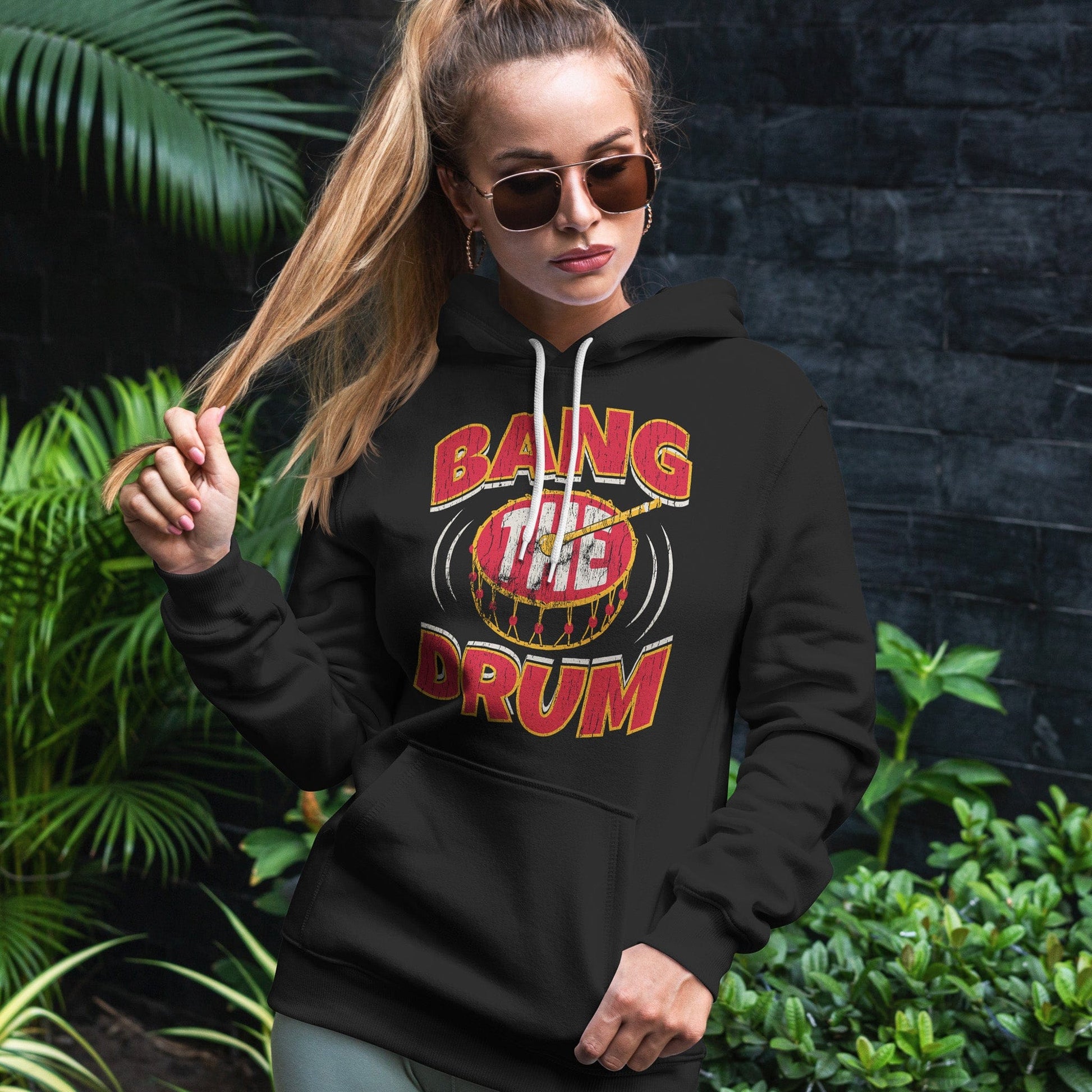 KC Swag | Kansas City Chiefs white/red/gold BAG THE DRUM on black sponge-fleece pullover hoodie worn by female model standing in front of a dark gray wall and green foliage
