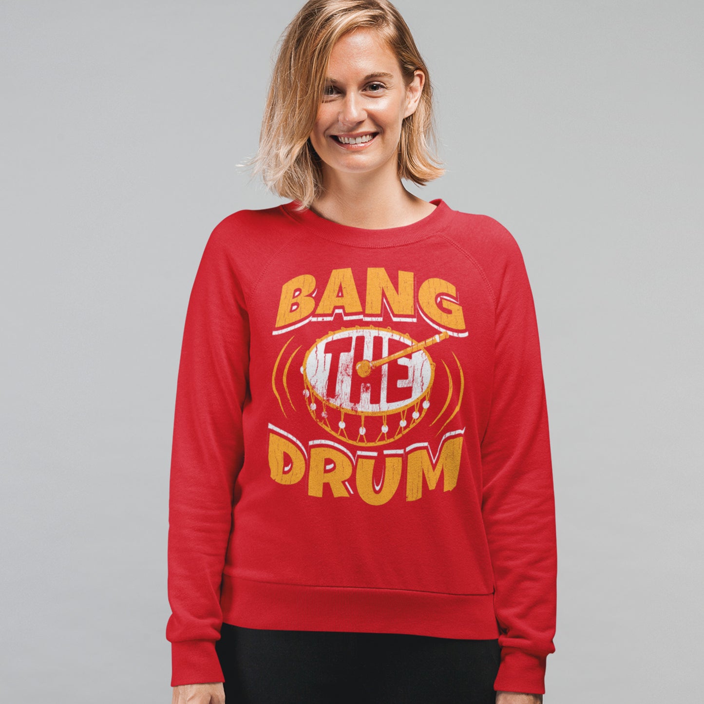 KC Swag Kansas City Chiefs Distressed White & Gold Band The Drum on a Red Crewneck Sweatshirt worn by a smiling female model standing in front of a gray studio wall