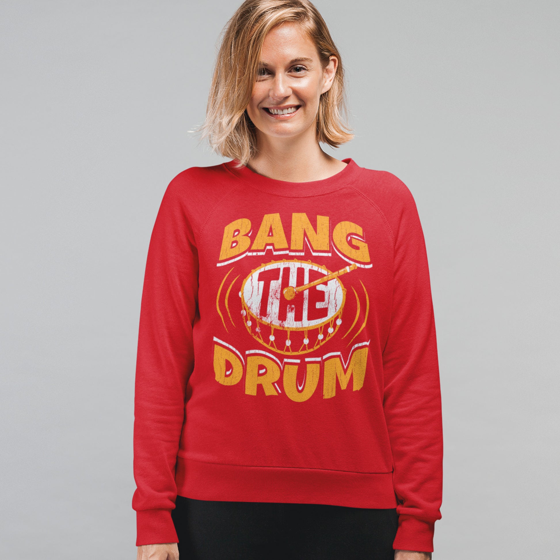 KC Swag Kansas City Chiefs Distressed White & Gold Band The Drum on a Red Crewneck Sweatshirt worn by a smiling female model standing in front of a gray studio wall