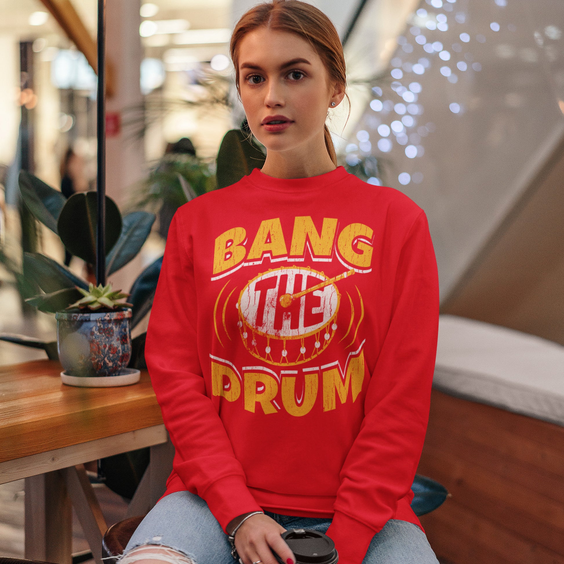 KC Swag Kansas City Chiefs Distressed White & Gold Band The Drum on a Red Crewneck Sweatshirt worn by female model sitting in a wood-tones coffee shop in a mall
