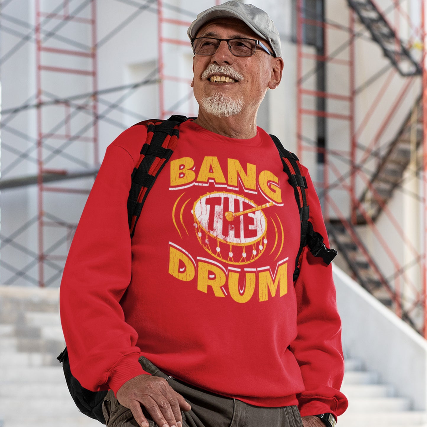 KC Swag Kansas City Chiefs Distressed White & Gold Band The Drum on a Red Crewneck Sweatshirt worn by an older male model wearing a hat and backpack standing on stairs in front of a construction scaffold