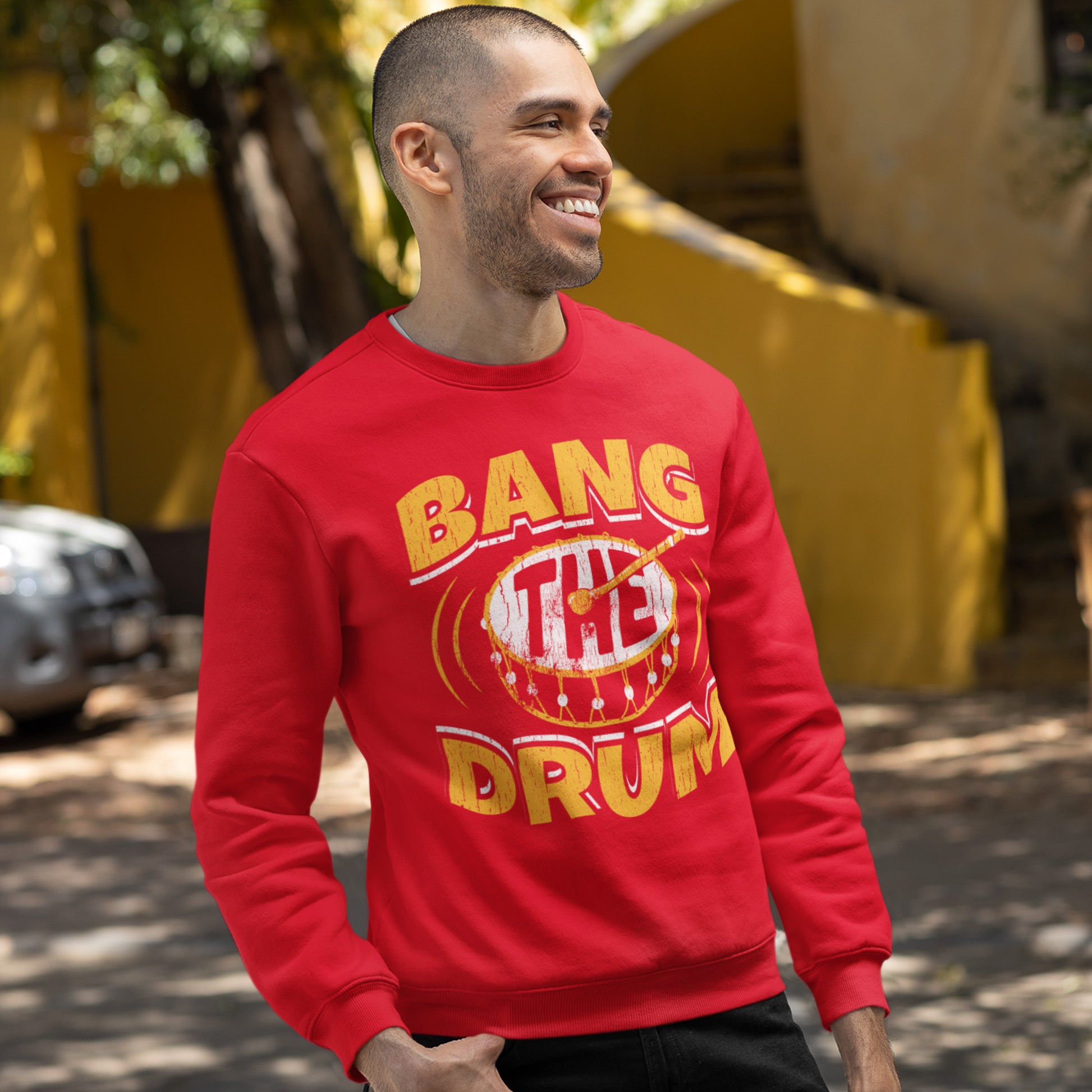 KC Swag Kansas City Chiefs Distressed White & Gold Band The Drum on a Red Crewneck Sweatshirt worn by a male model in a parking lot in front of a yellow concrete staircase