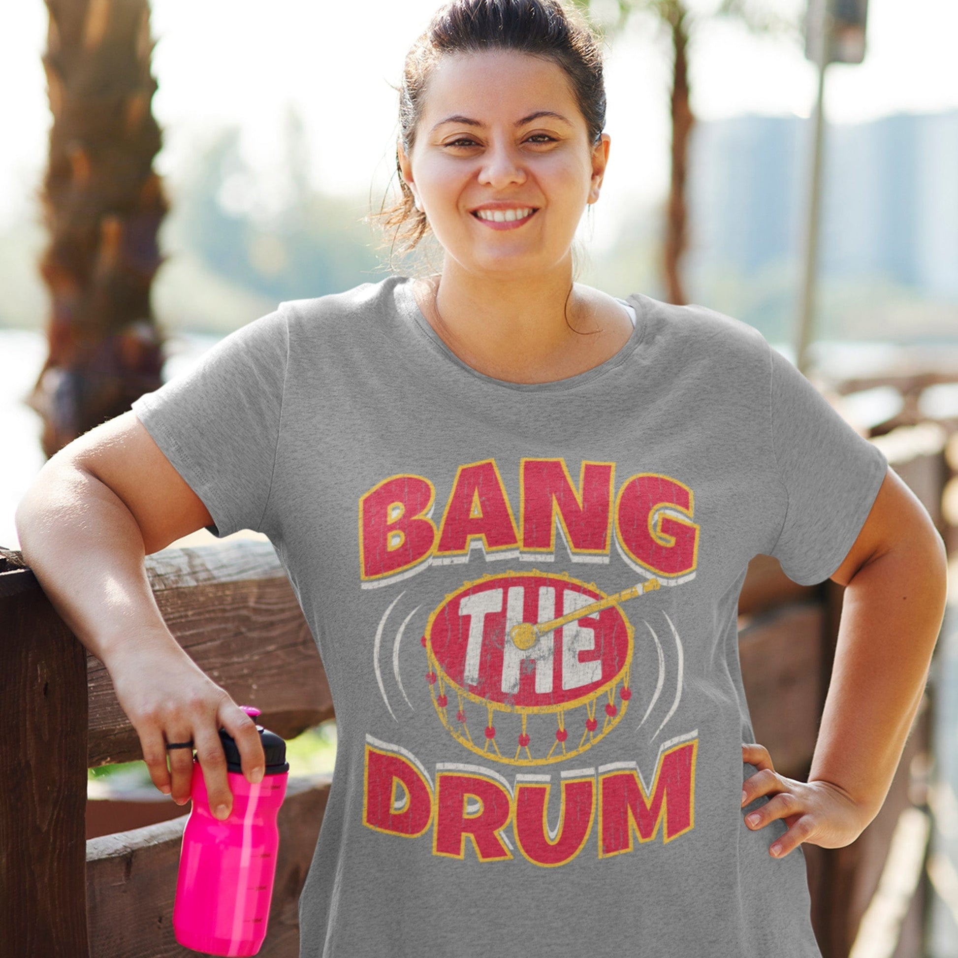 KC Swag | Kansas City Chiefs white, gold & red BANG THE DRUM on athletic heather gray unisex t-shirt worn by female model holding water bottle leaning on wood fence in a public park