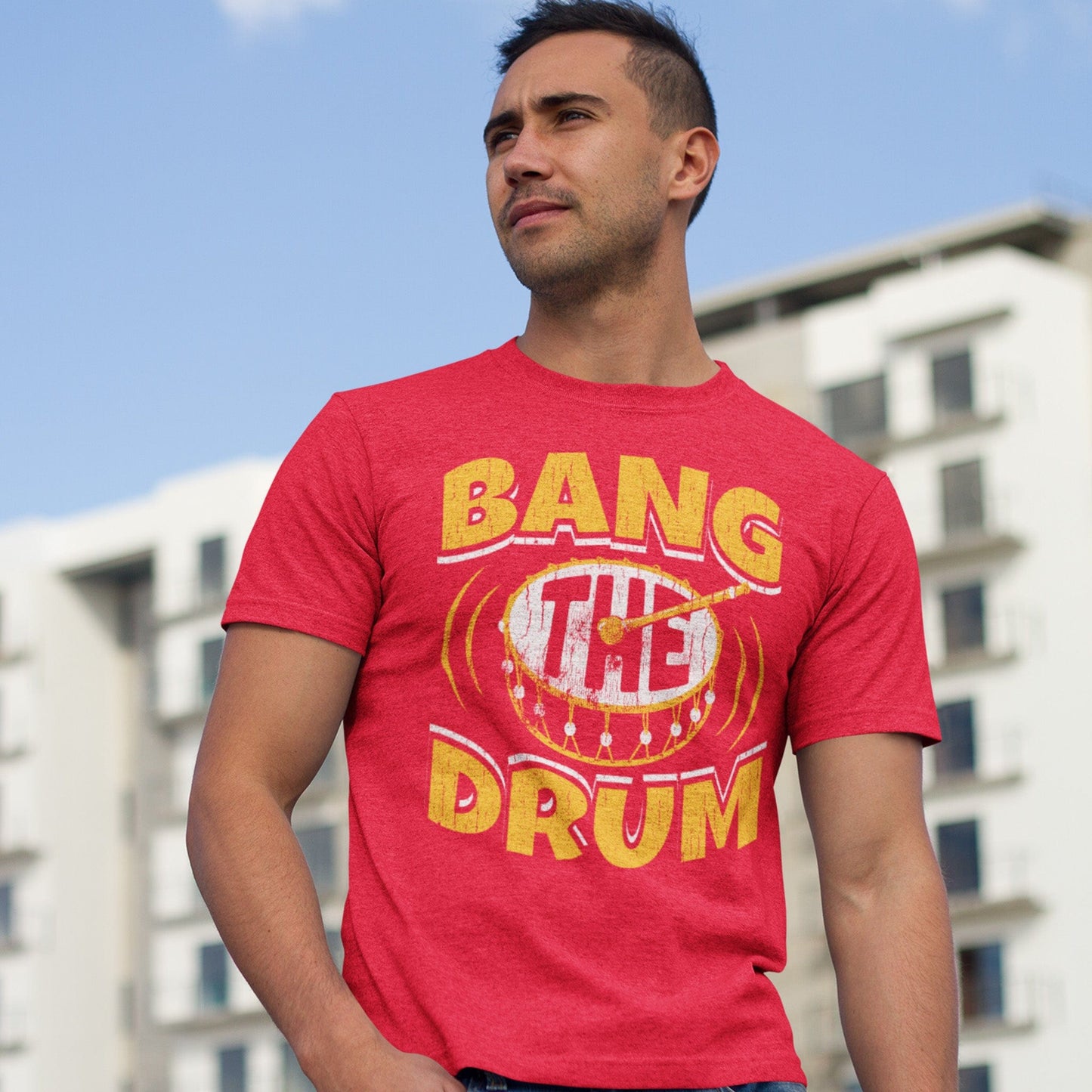 KC Swag | Kansas City Chiefs white & gold BANG THE DRUM on heather red unisex t-shirt worn by male model standing in front of a tall apartment building