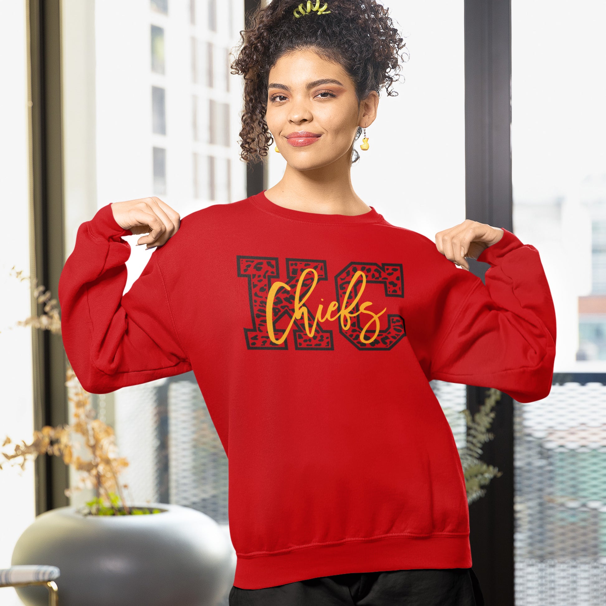 KC Swag Kansas City Chiefs Black & Gold Cheetah KC on a Red Crewneck Sweatshirt worn by female model pointing at her chest in a high-rise apartment