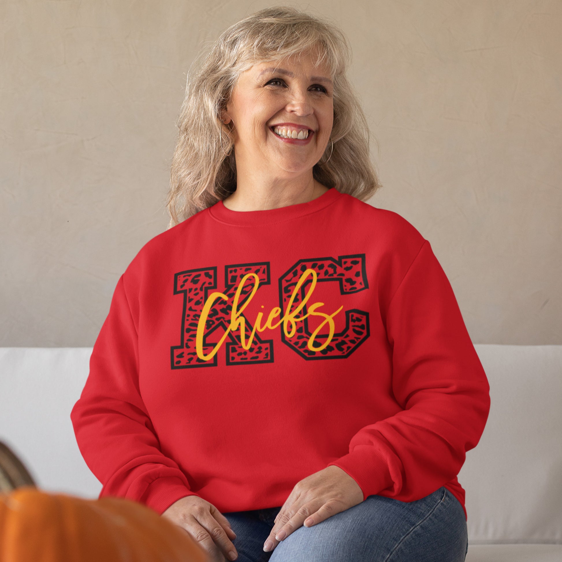 KC Swag Kansas City Chiefs Black & Gold Cheetah KC on a Red Crewneck Sweatshirt worn by an older female model sitting on a white couch in front of a beige wall