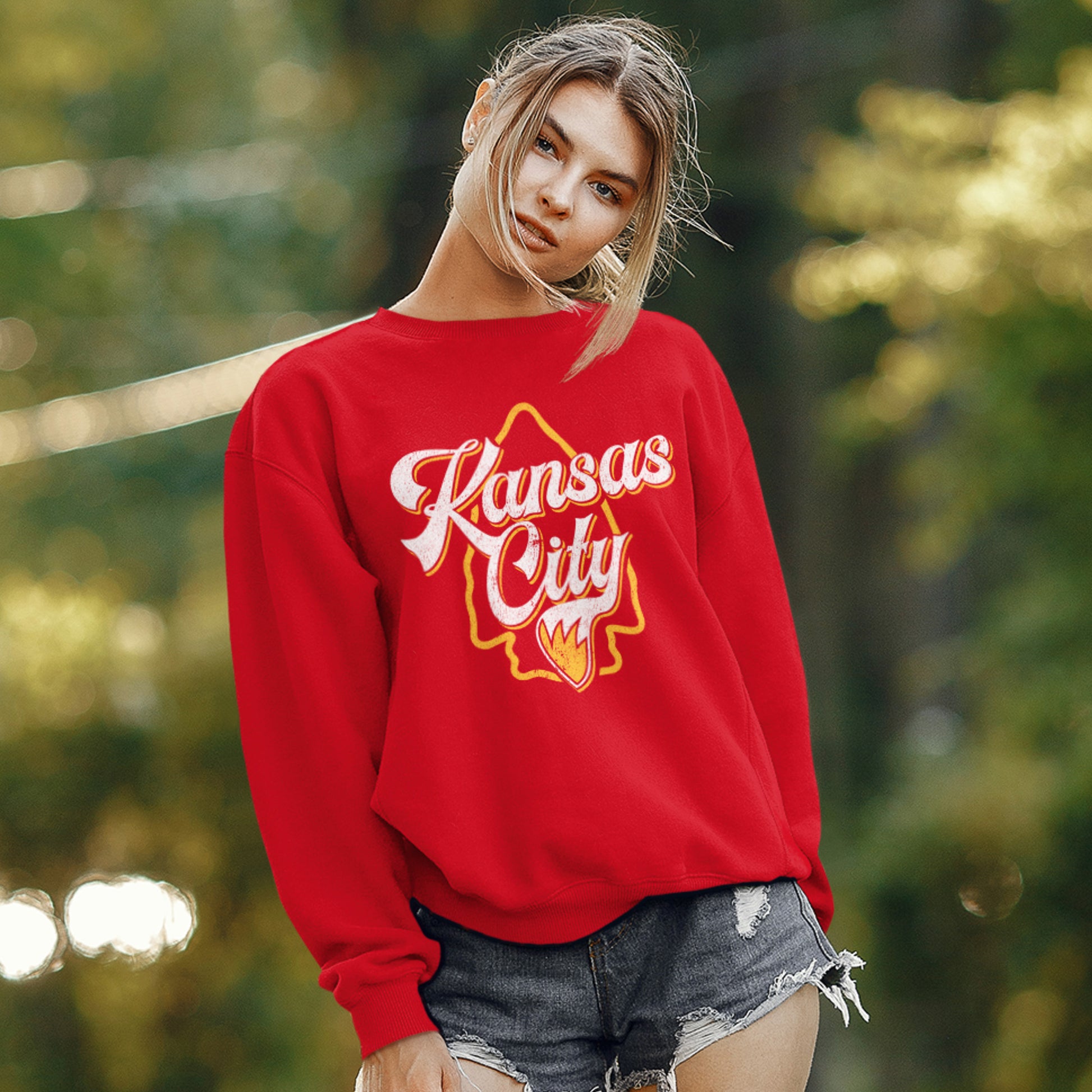 KC Swag Kansas City Chiefs Distressed White & Gold Wolftail KC on a Red Crewneck Sweatshirt worn by female wearing ripped jean-shorts standing in a public park