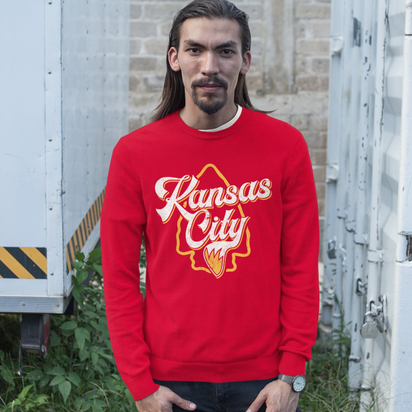 KC Swag Kansas City Chiefs Distressed White & Gold Wolftail KC on a Red Crewneck Sweatshirt worn by a skinny male model standing in a field near white shipping containers