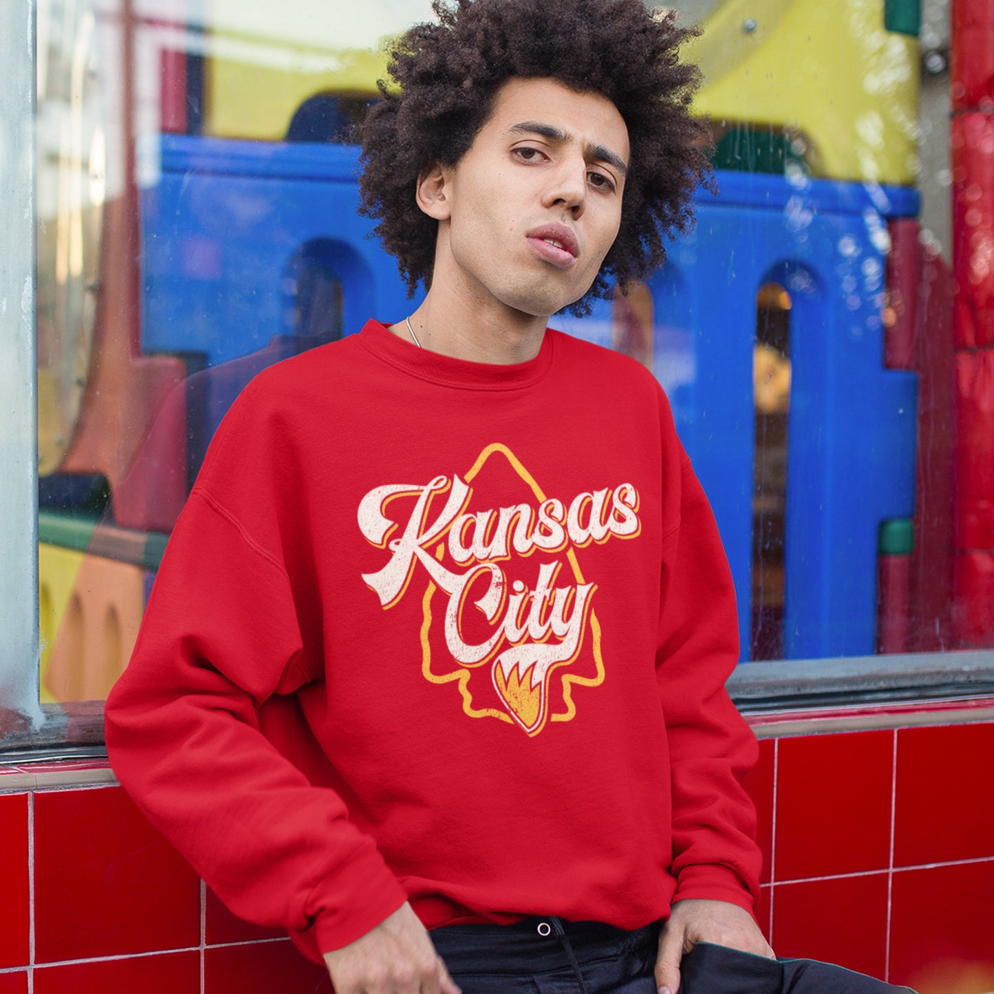 KC Swag Kansas City Chiefs Distressed White & Gold Wolftail KC on a Red Crewneck Sweatshirt worn by male model with an afro leaning against a storefront window